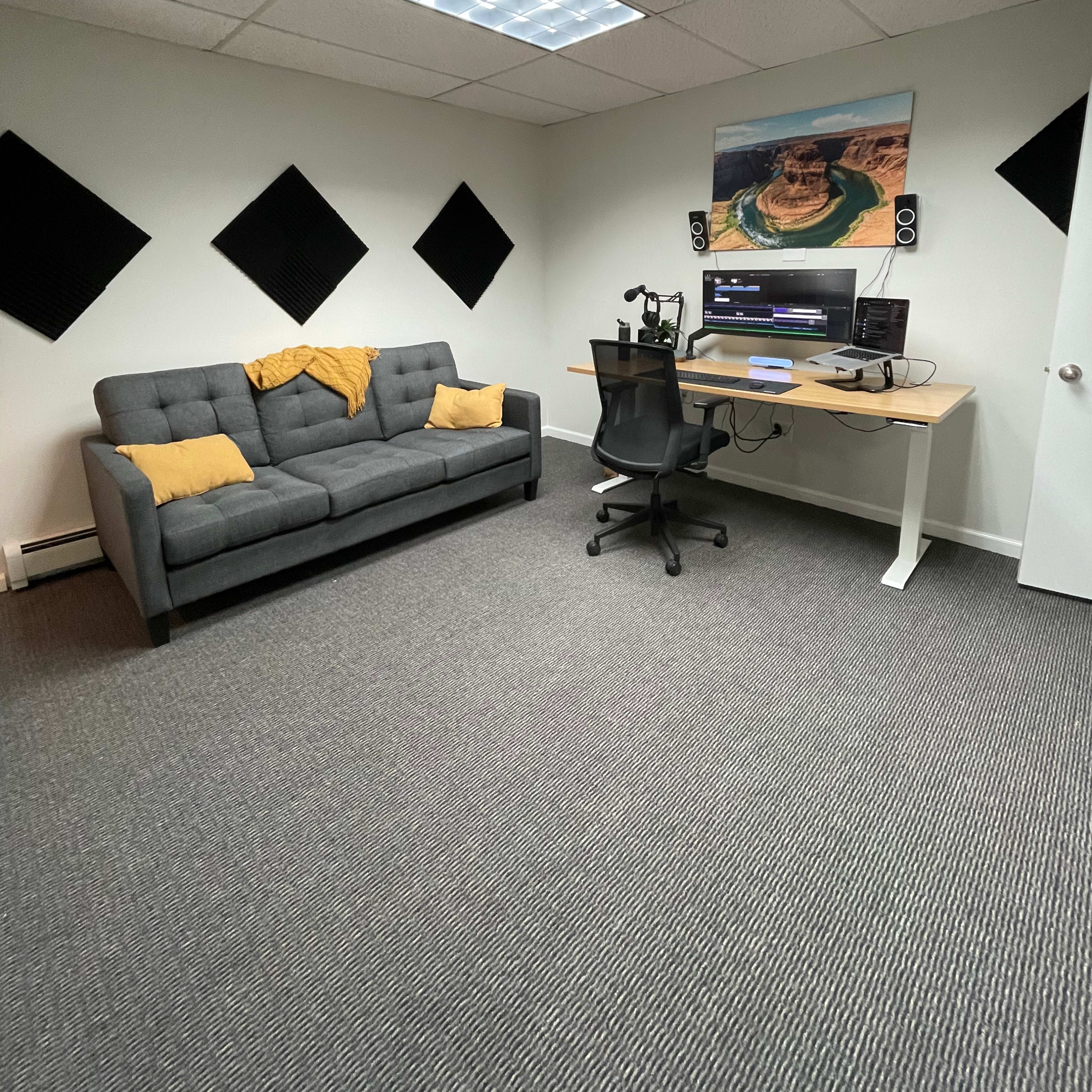 A small office space featuring a gray sofa with yellow throw pillows, a desk with audio equipment, and soundproofing panels on the walls.