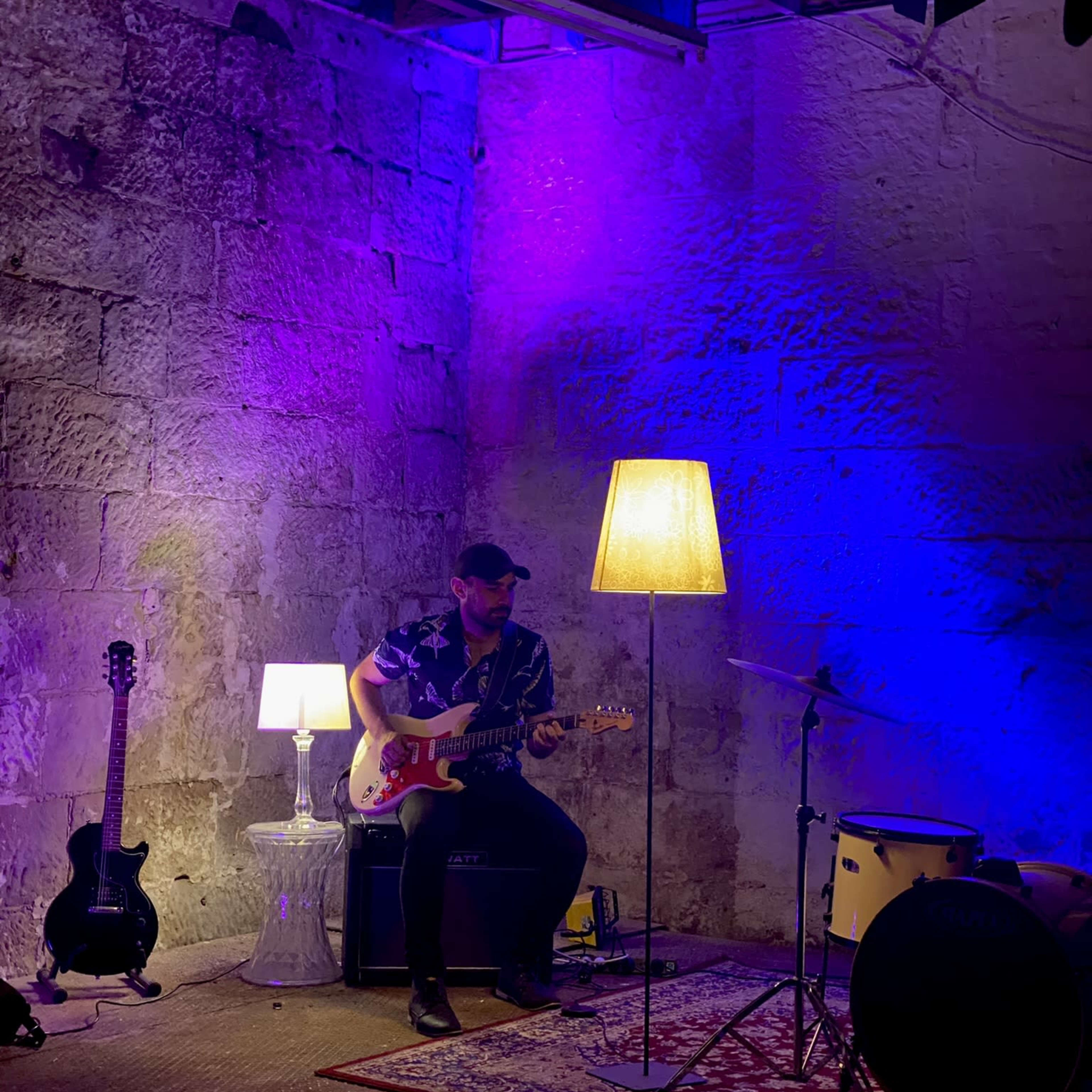 A musician sits on a stool playing an electric guitar in a dimly lit space with purple and blue lighting, accompanied by a lamp and a drum set.
