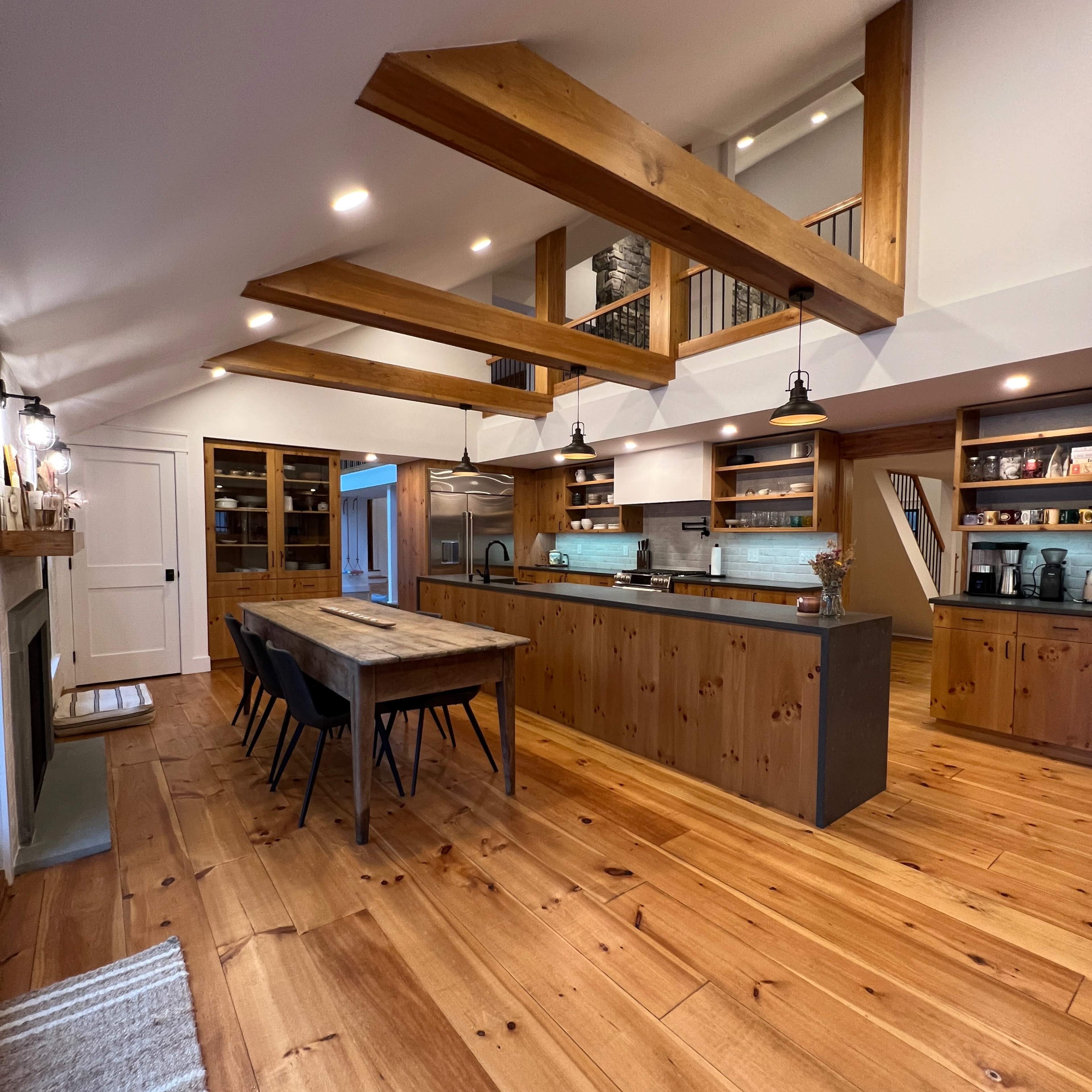 A modern kitchen featuring a large wooden island, a dining table with black chairs, and wooden beams supporting the ceiling.