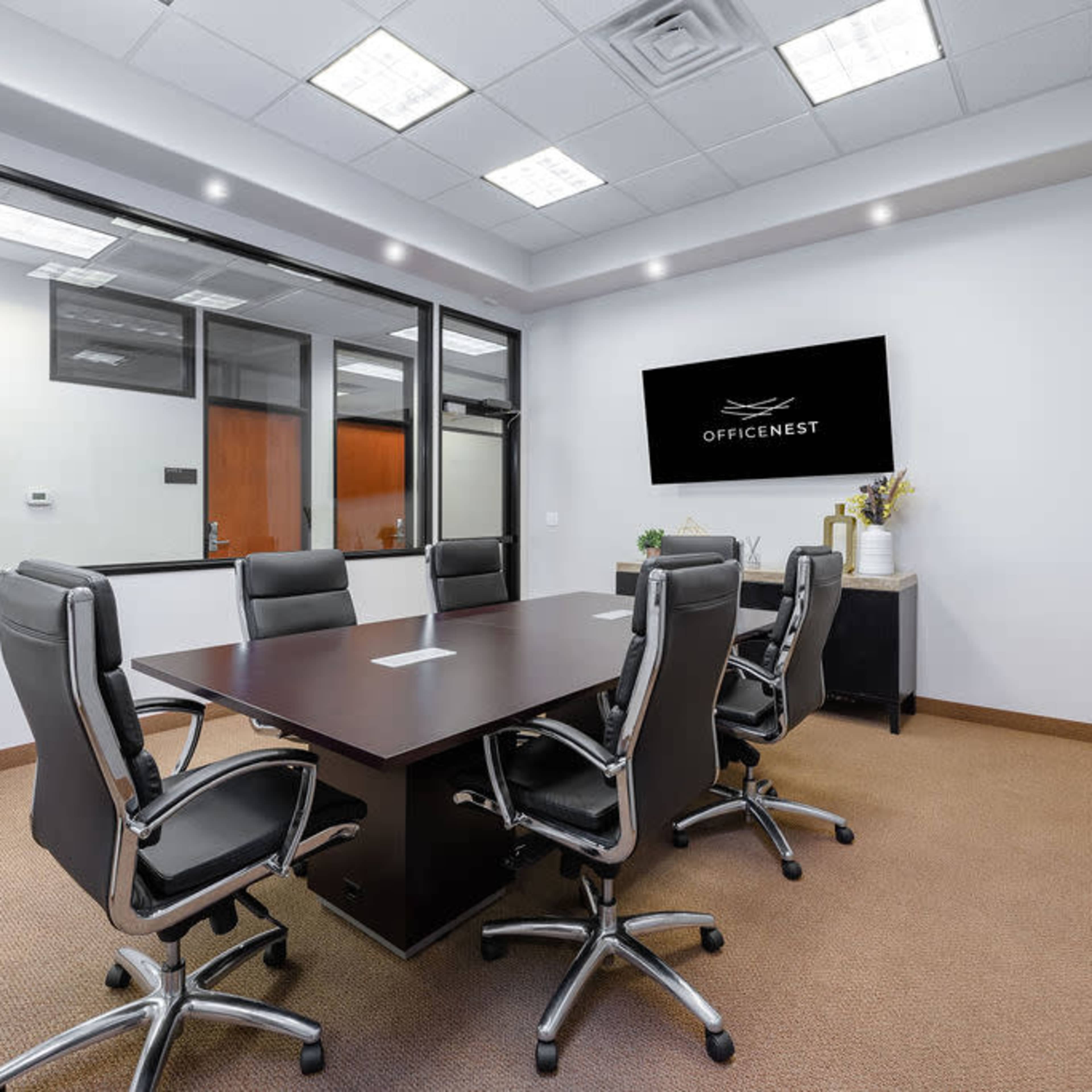 A modern conference room features a large dark table surrounded by six black ergonomic chairs, with a wall-mounted television displaying the logo "OFFICENEST."