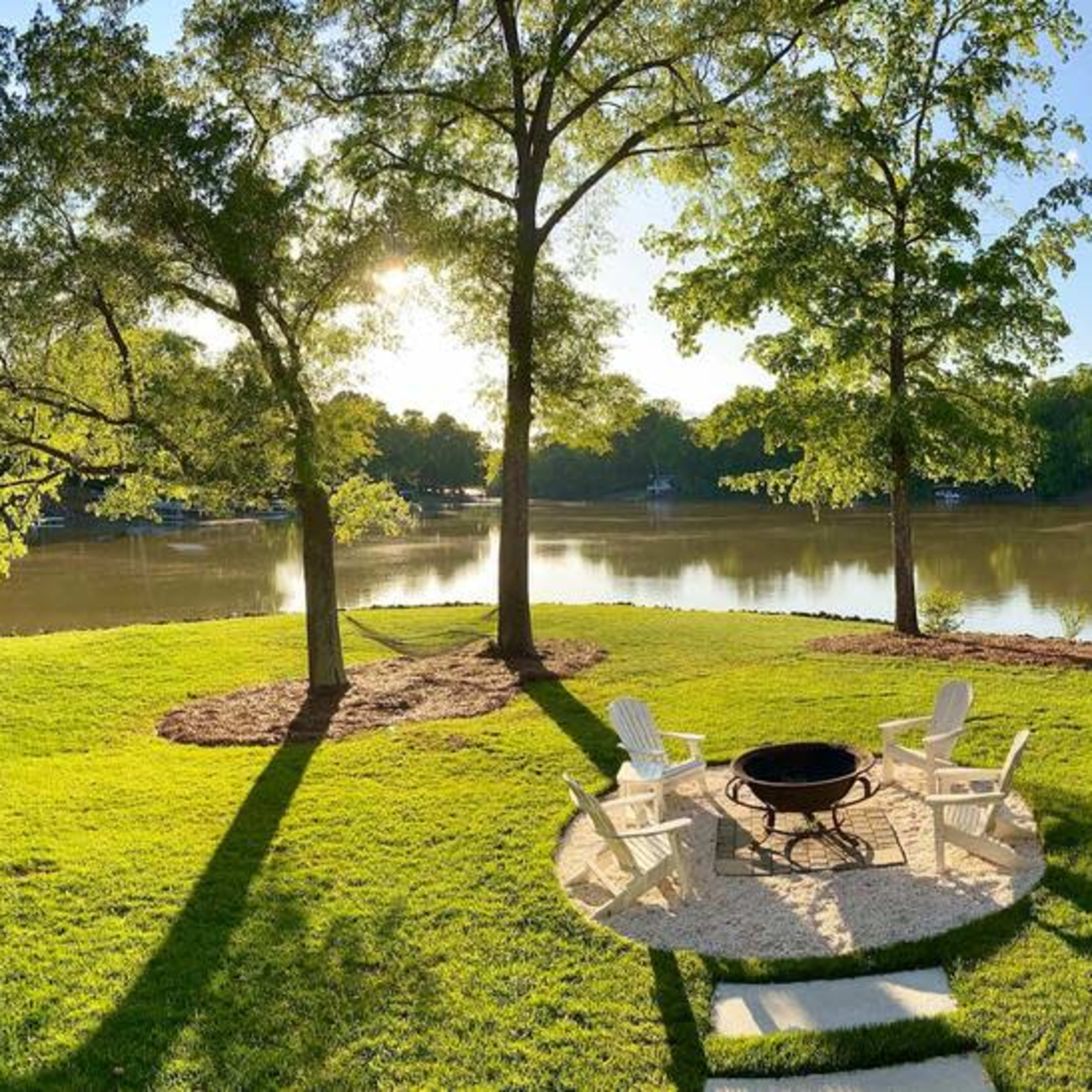 A landscaped yard by a calm lake, featuring a fire pit surrounded by chairs and bordered by trees.