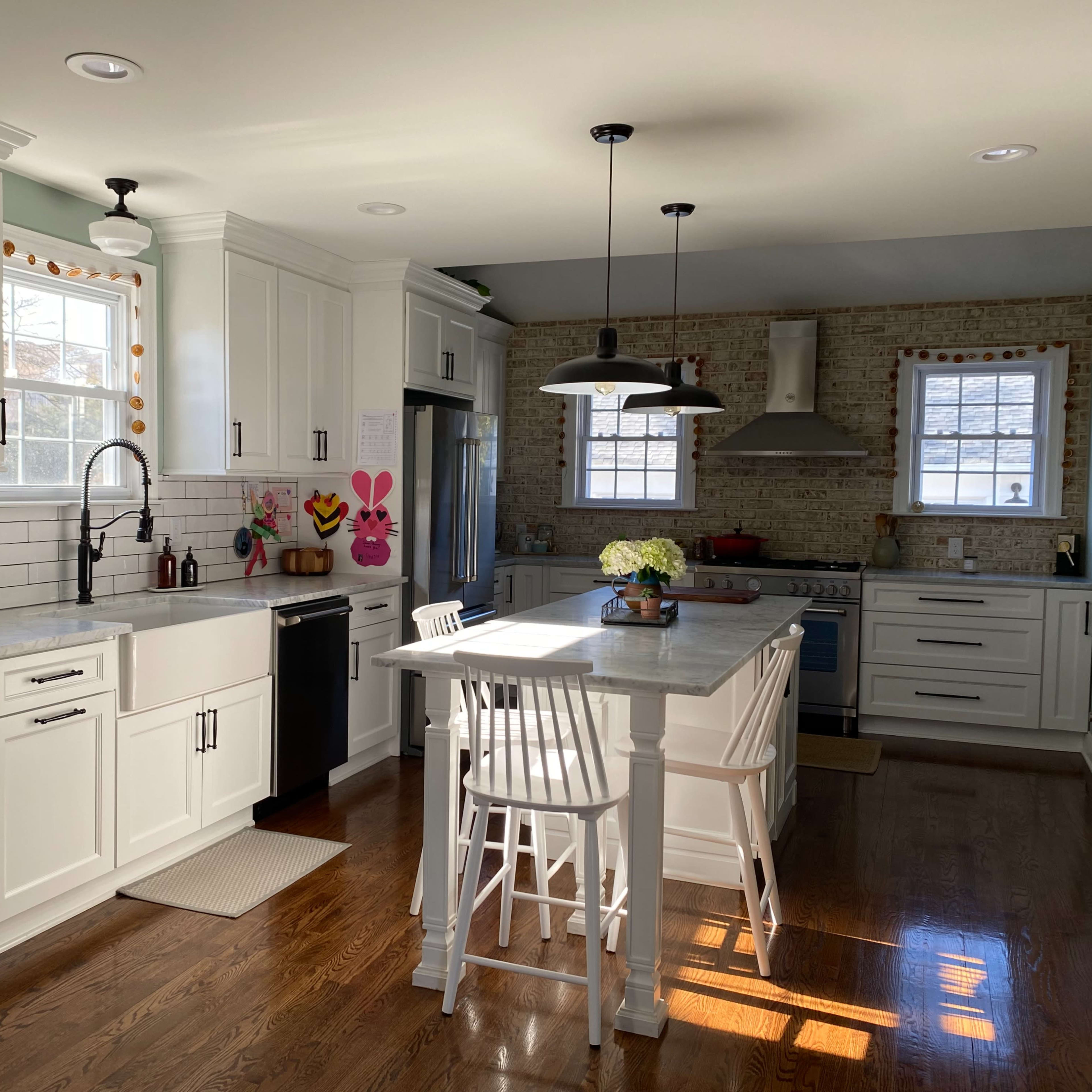 A bright kitchen features white cabinets, a central island with stools, and brick walls, illuminated by natural light from two windows.