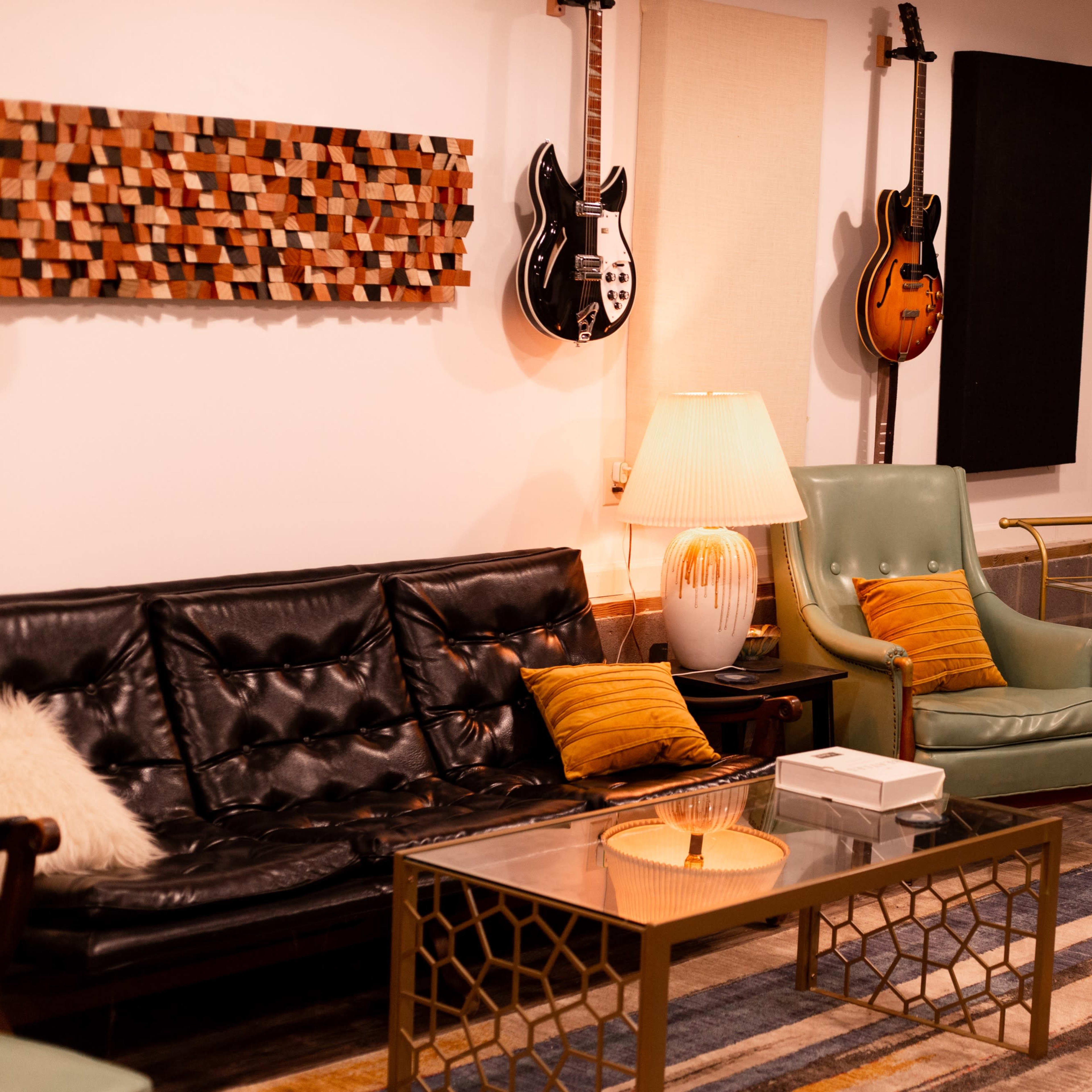 A cozy music studio featuring a black leather sofa, a green armchair, a coffee table, and various guitars displayed on the walls.