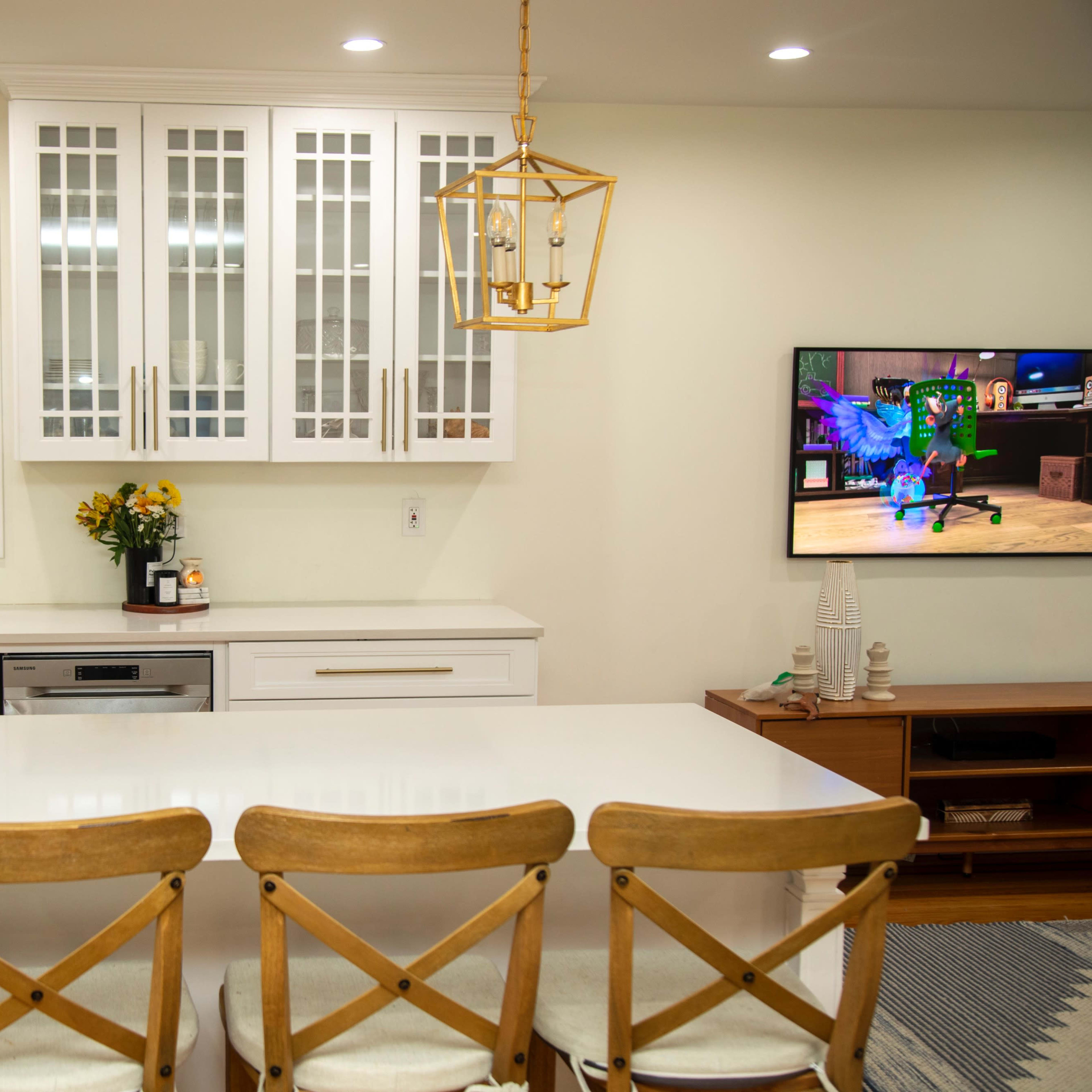 A modern kitchen features a central island with wooden chairs, white cabinetry with glass doors, and a television mounted on the wall displaying a colorful image.