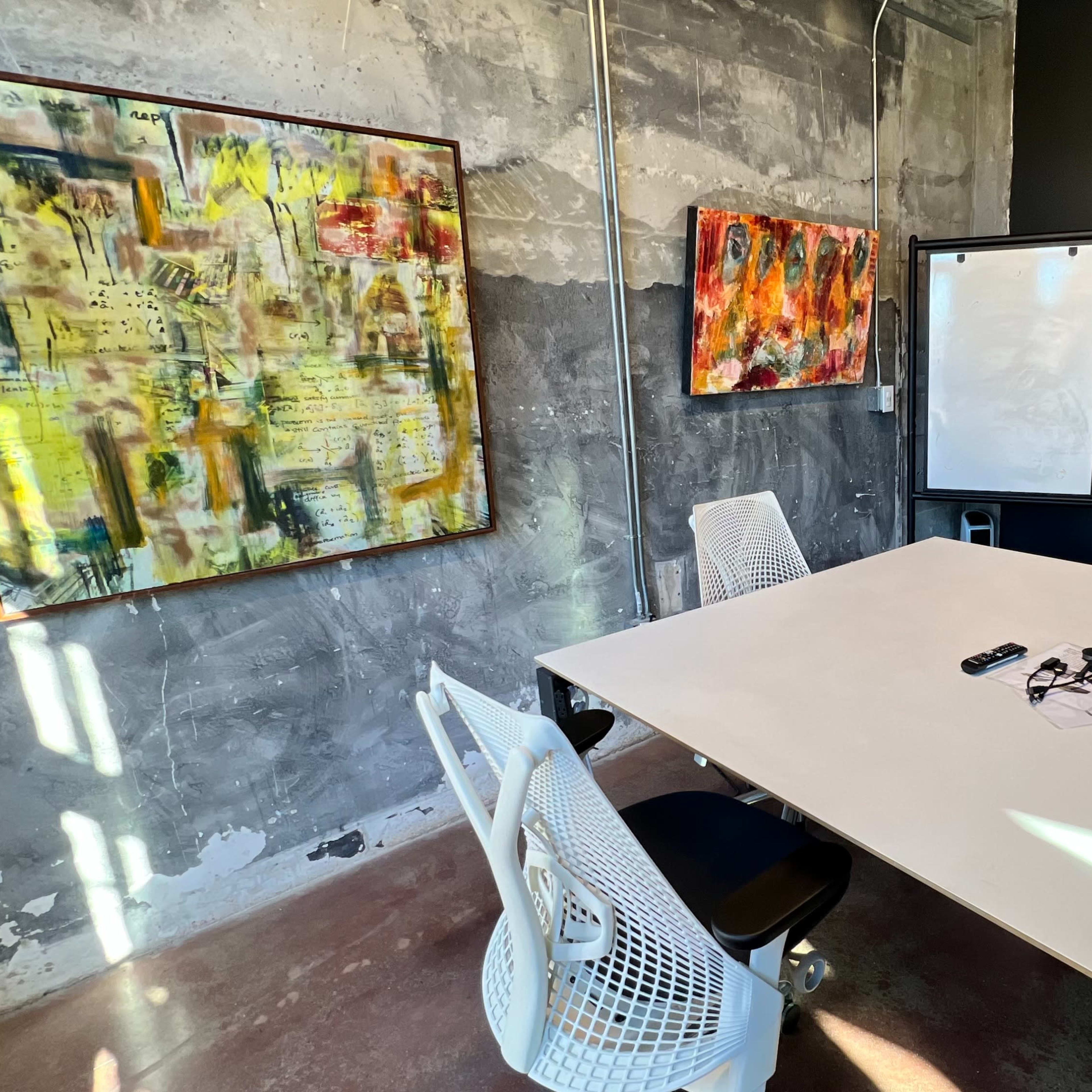 A modern meeting room features a white table surrounded by four white chairs, with abstract artwork on the walls and a wall-mounted screen.