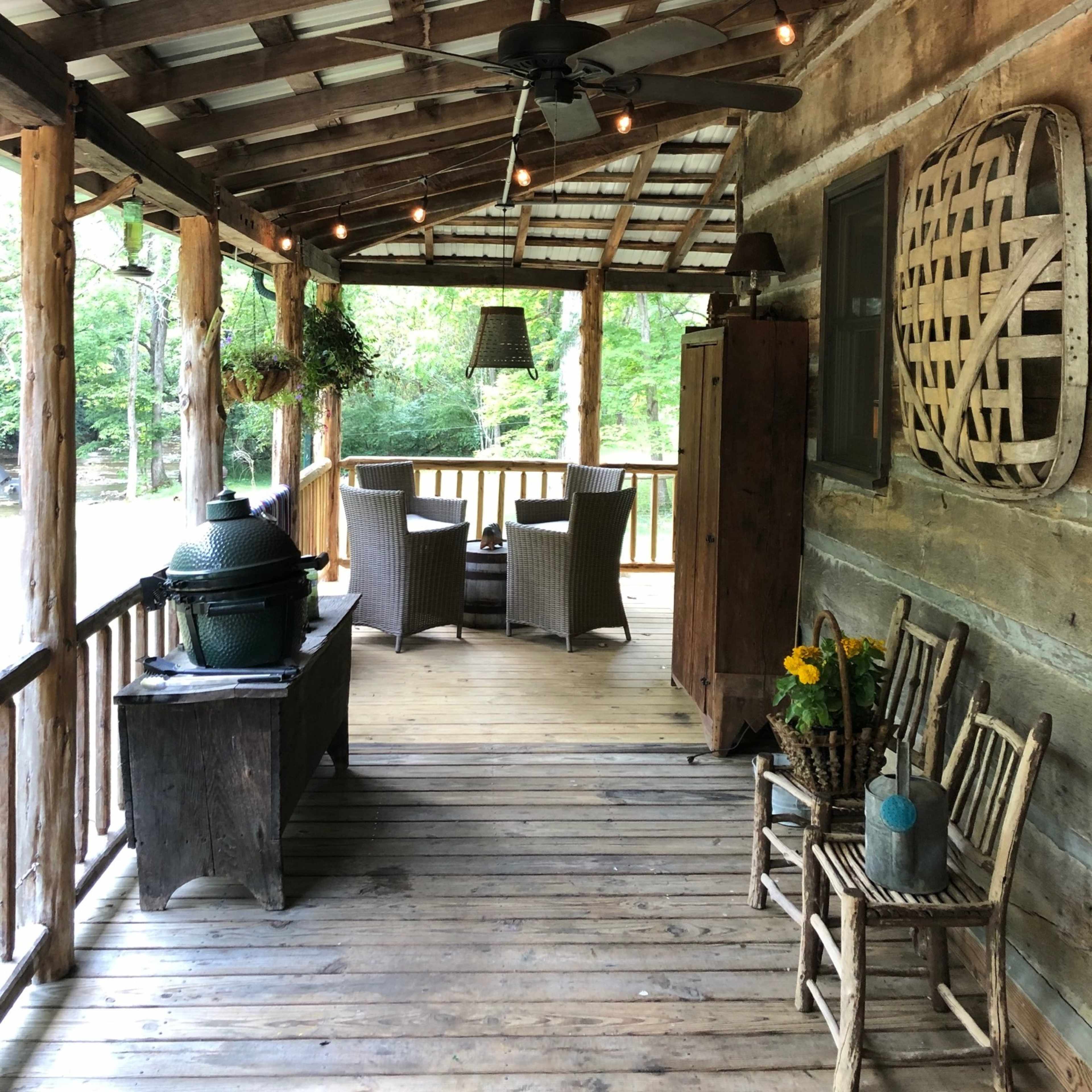 A rustic porch with wooden beams, a ceiling fan, and string lights, furnished with wicker chairs and a table, surrounded by greenery.