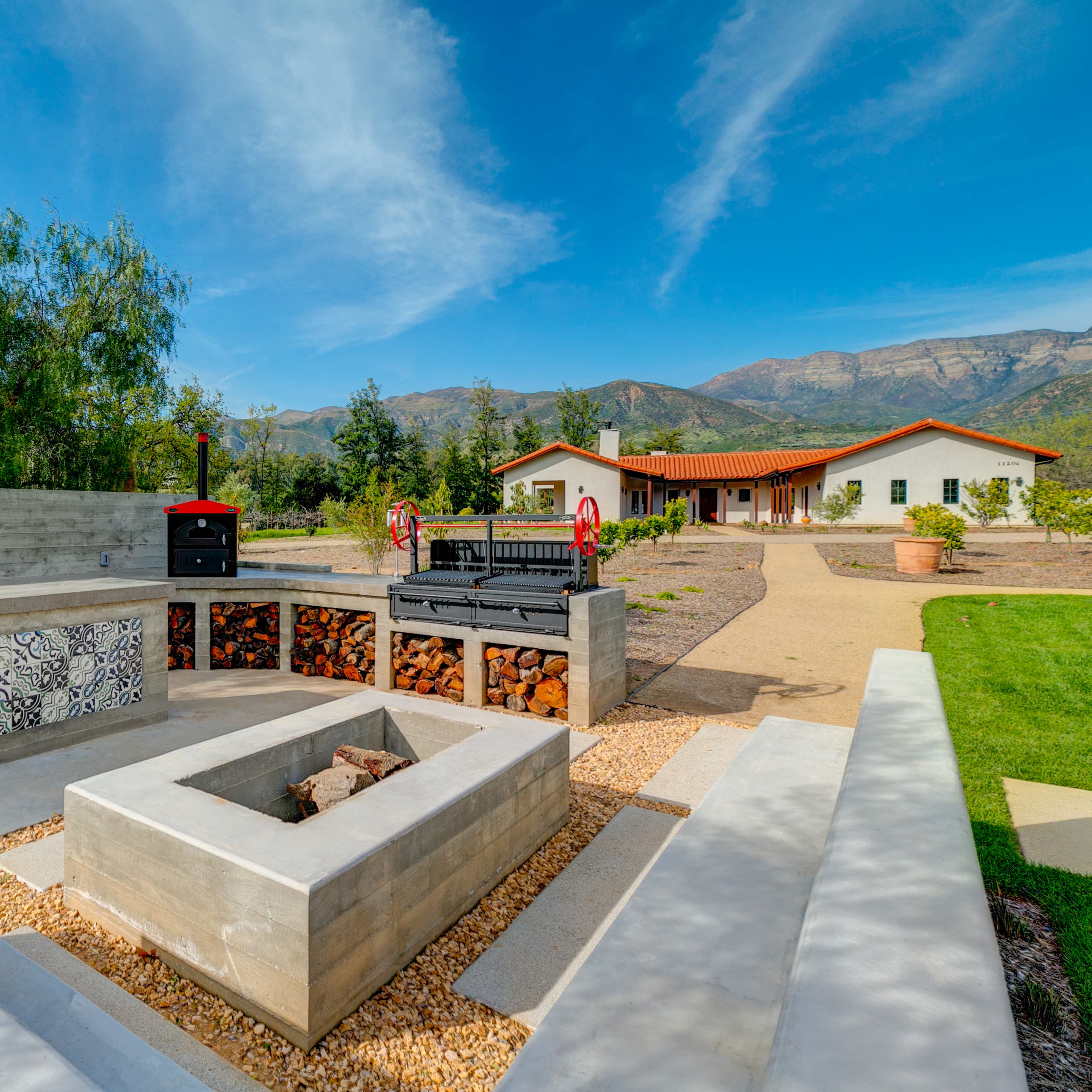 A modern outdoor kitchen area with a stone fire pit and a grill, surrounded by landscaped grounds and mountains in the background.