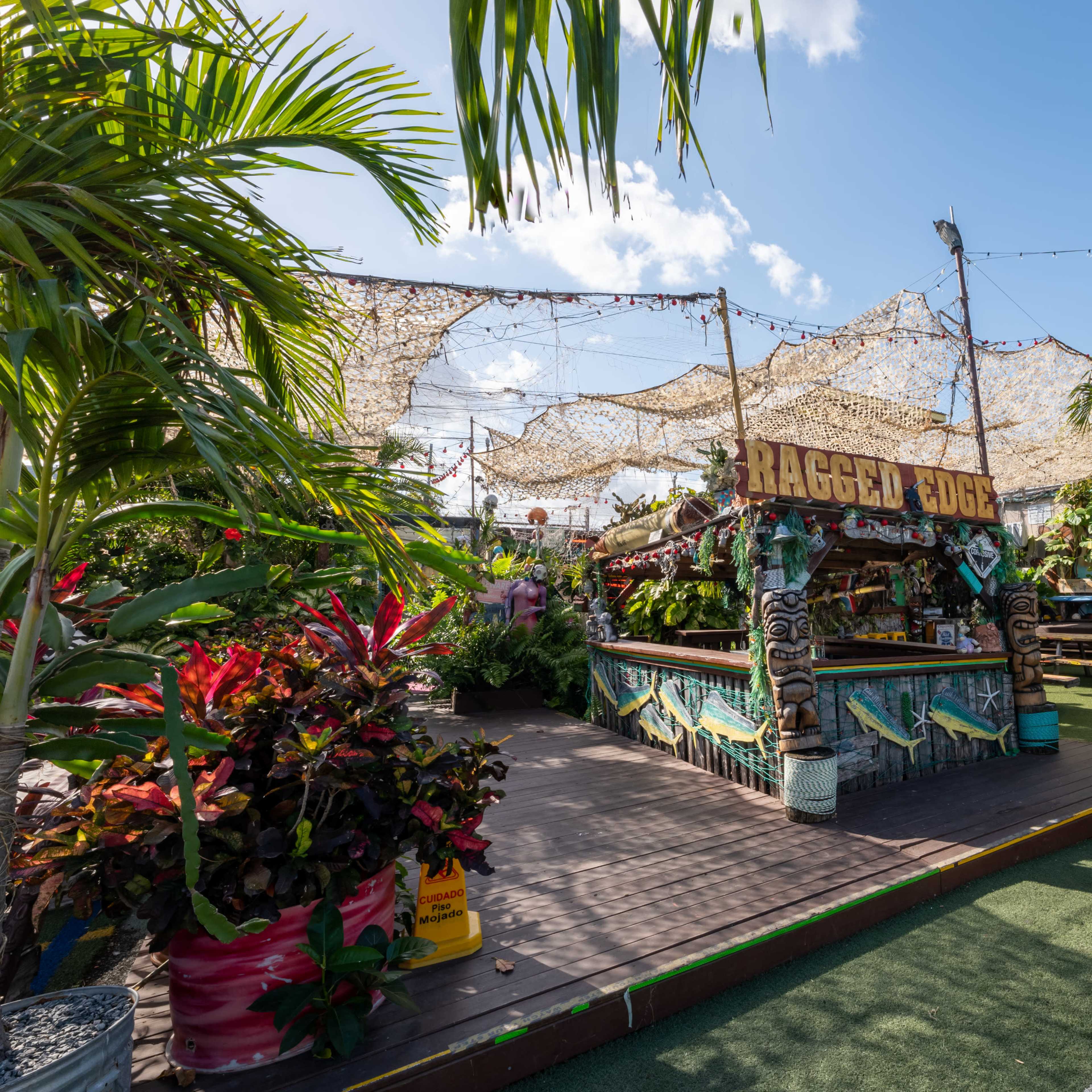 A colorful outdoor bar named "Ragged Edge" is surrounded by tropical plants and decorated with fishing nets and nautical elements.