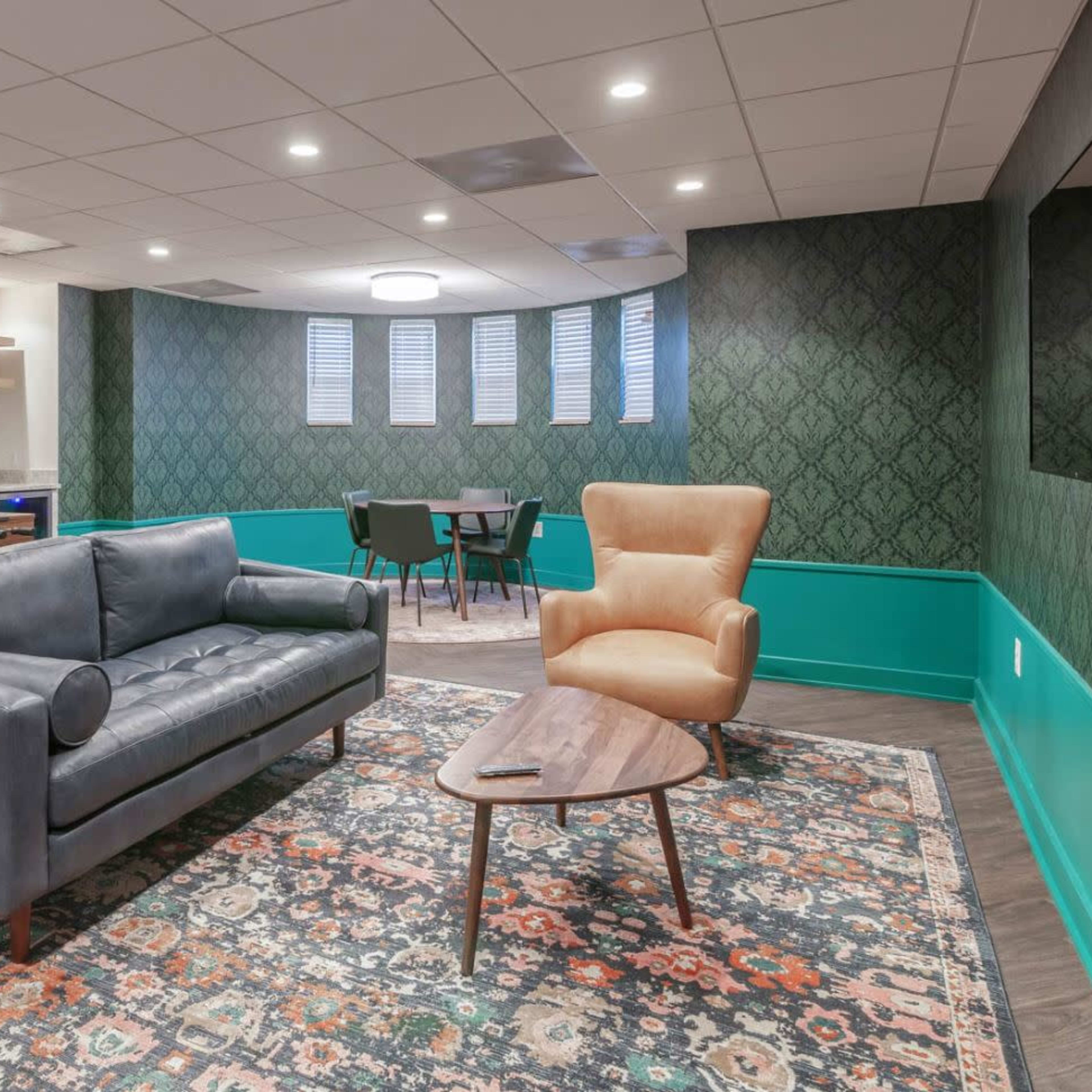 A modern lounge area featuring a dark couch, a light brown armchair, a coffee table, and a foosball table, all set against patterned wallpaper and teal accents.
