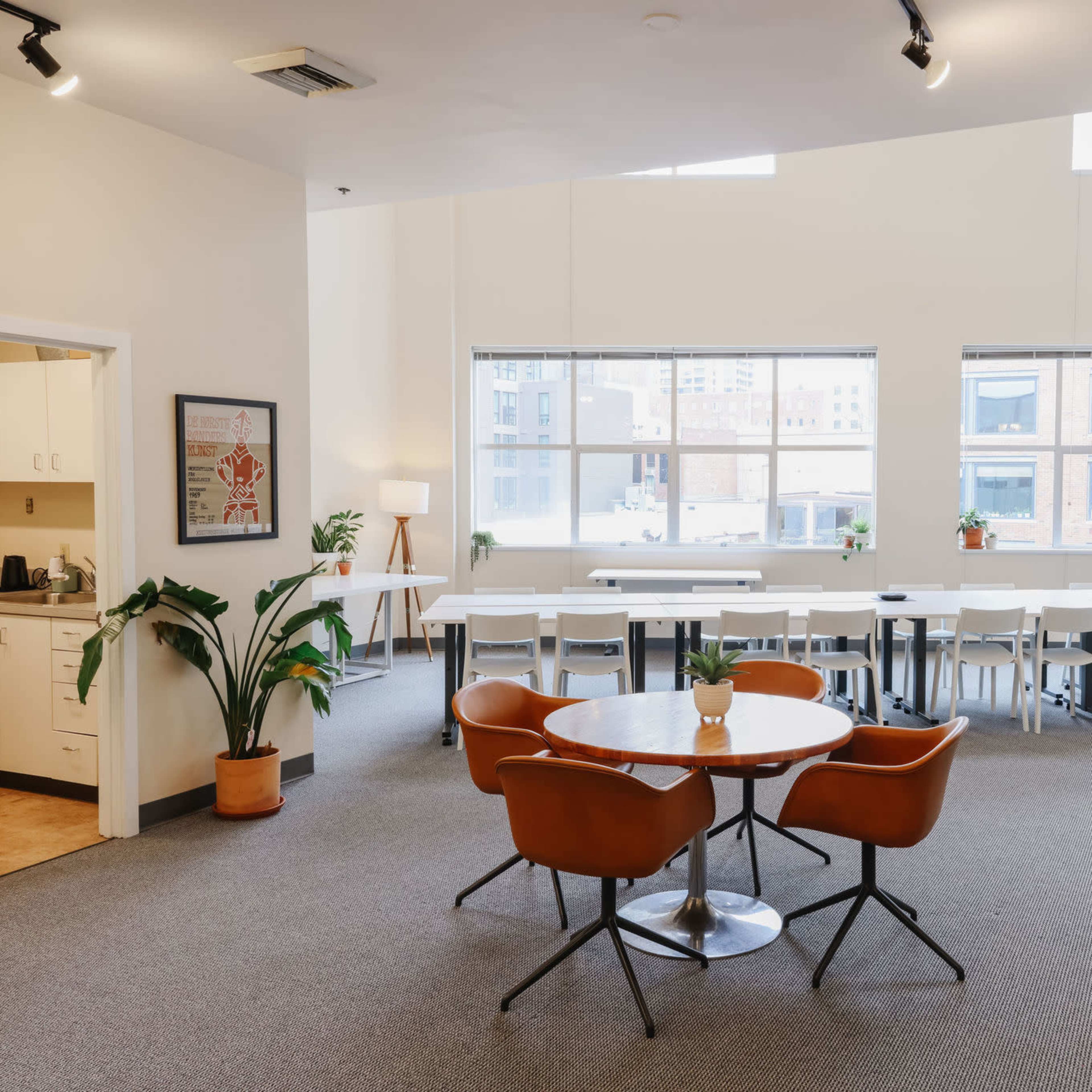 A spacious, modern communal area with a round table and chairs, large windows, and a kitchenette in the background.