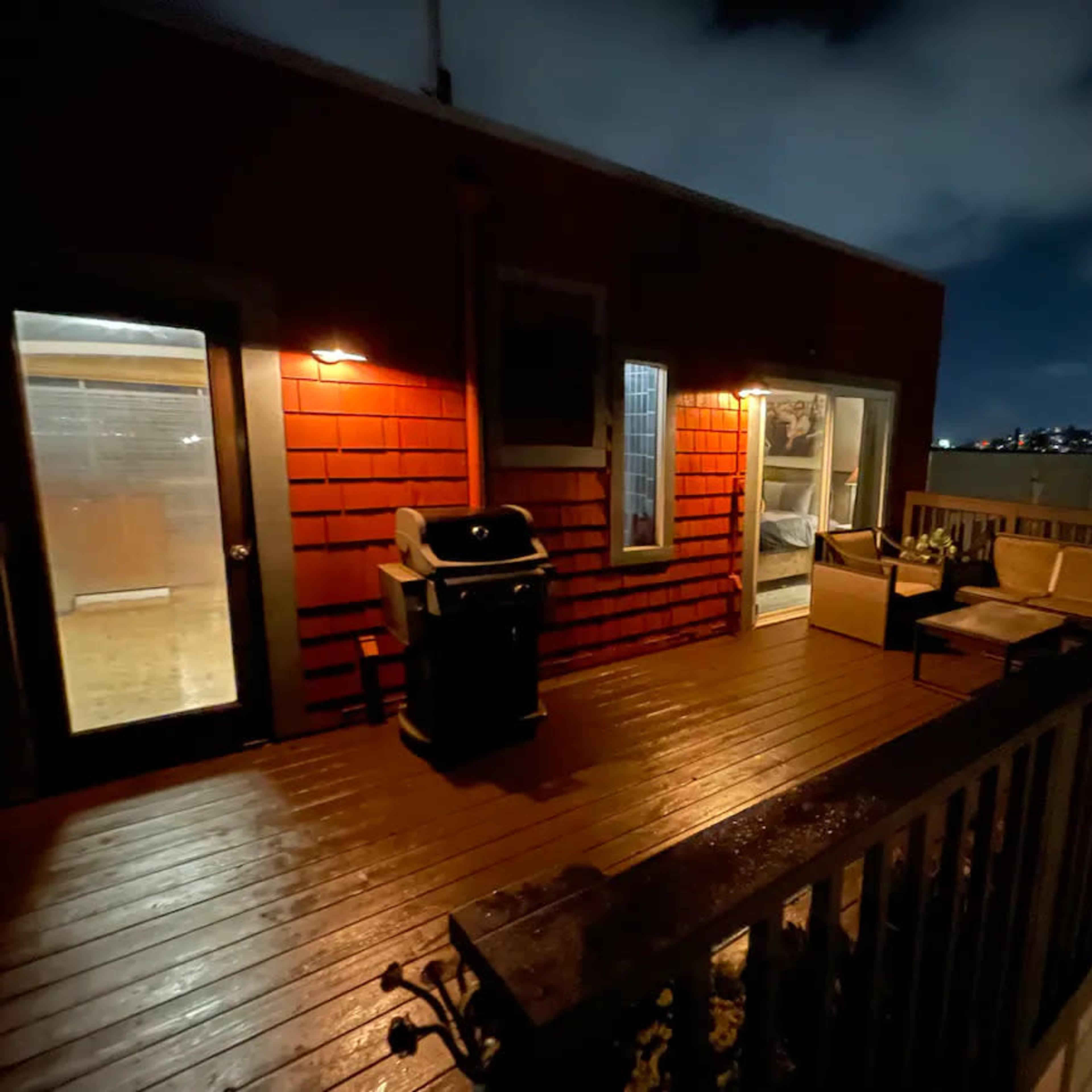 A wooden deck with outdoor seating and a barbecue grill is illuminated at night, overlooking a city skyline.