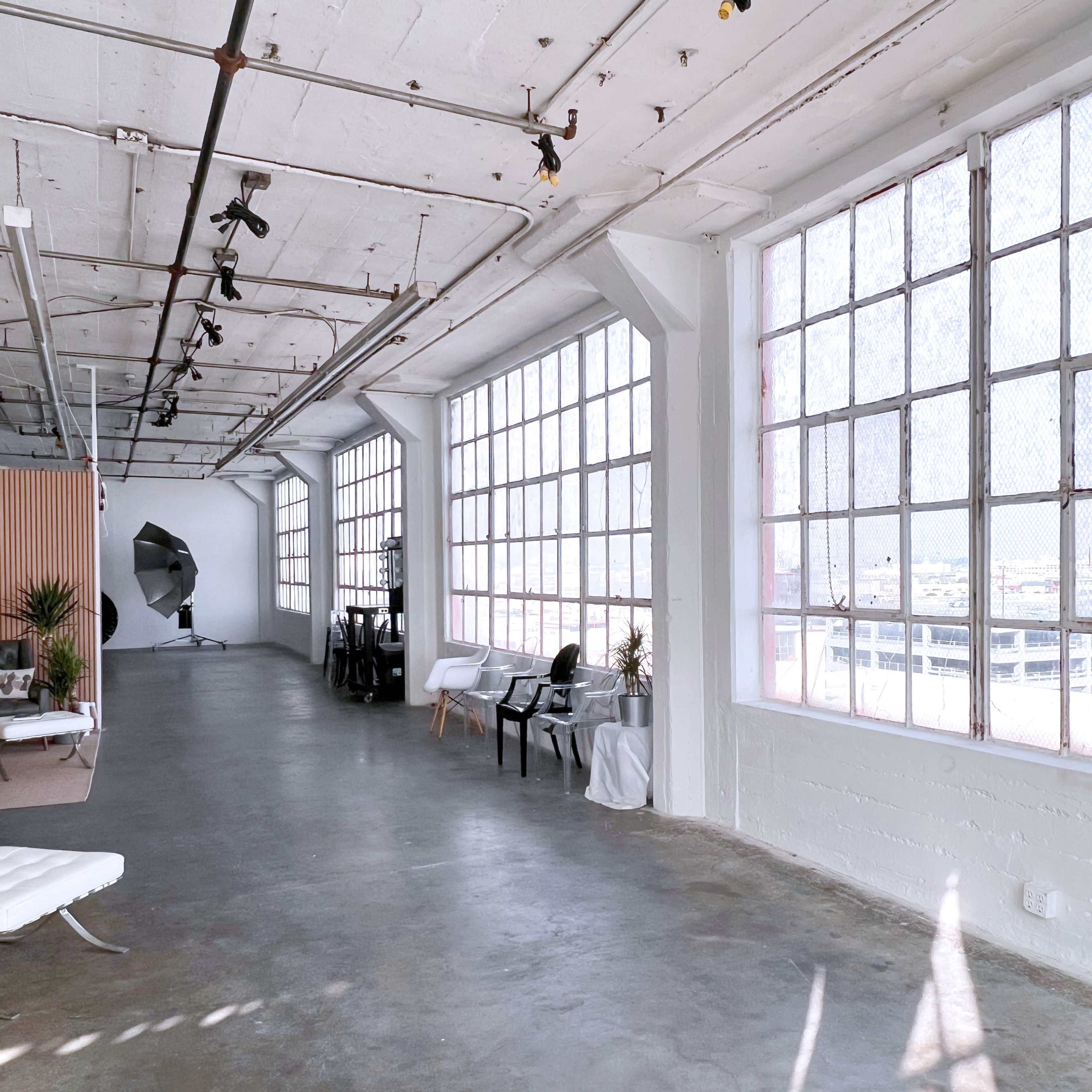 A spacious, well-lit industrial room featuring large windows, concrete flooring, and various chairs arranged along the sides.