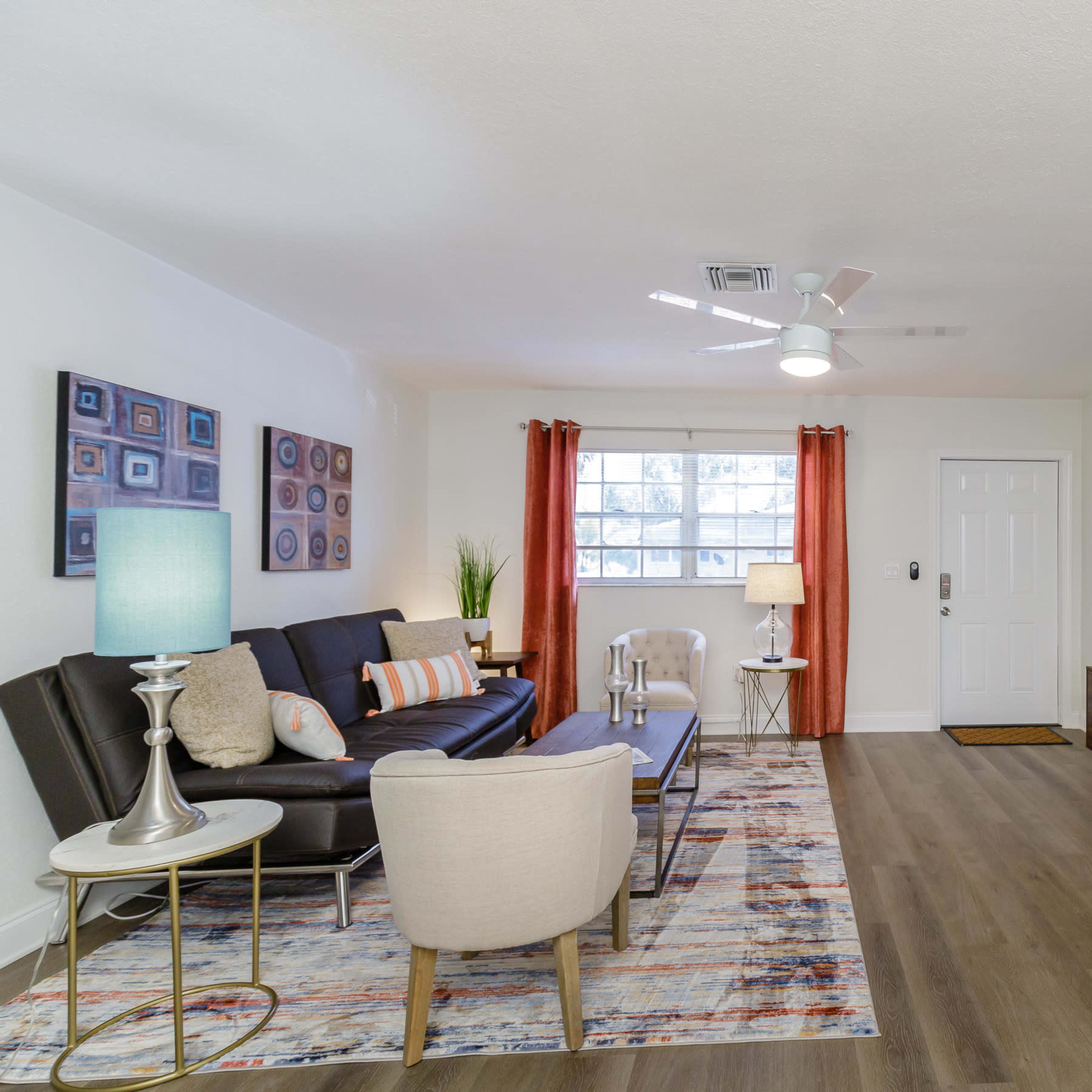 A modern living room features a black sofa, a blue accent chair, a coffee table, and a large television mounted on the wall, with decorative artwork and curtains framing the windows.