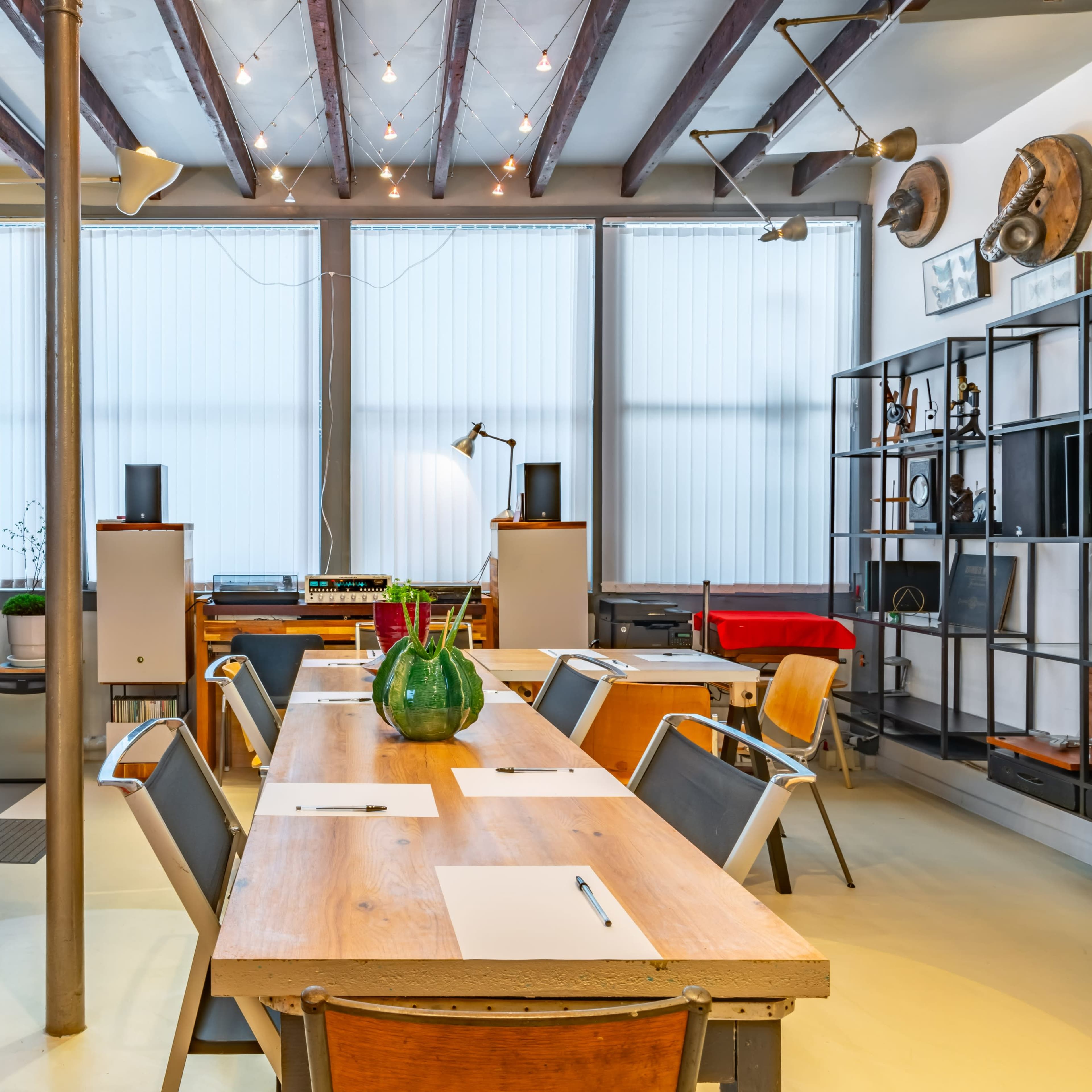 A modern conference room features a long wooden table surrounded by chairs, with large windows, metal shelves, and lockers in the background.