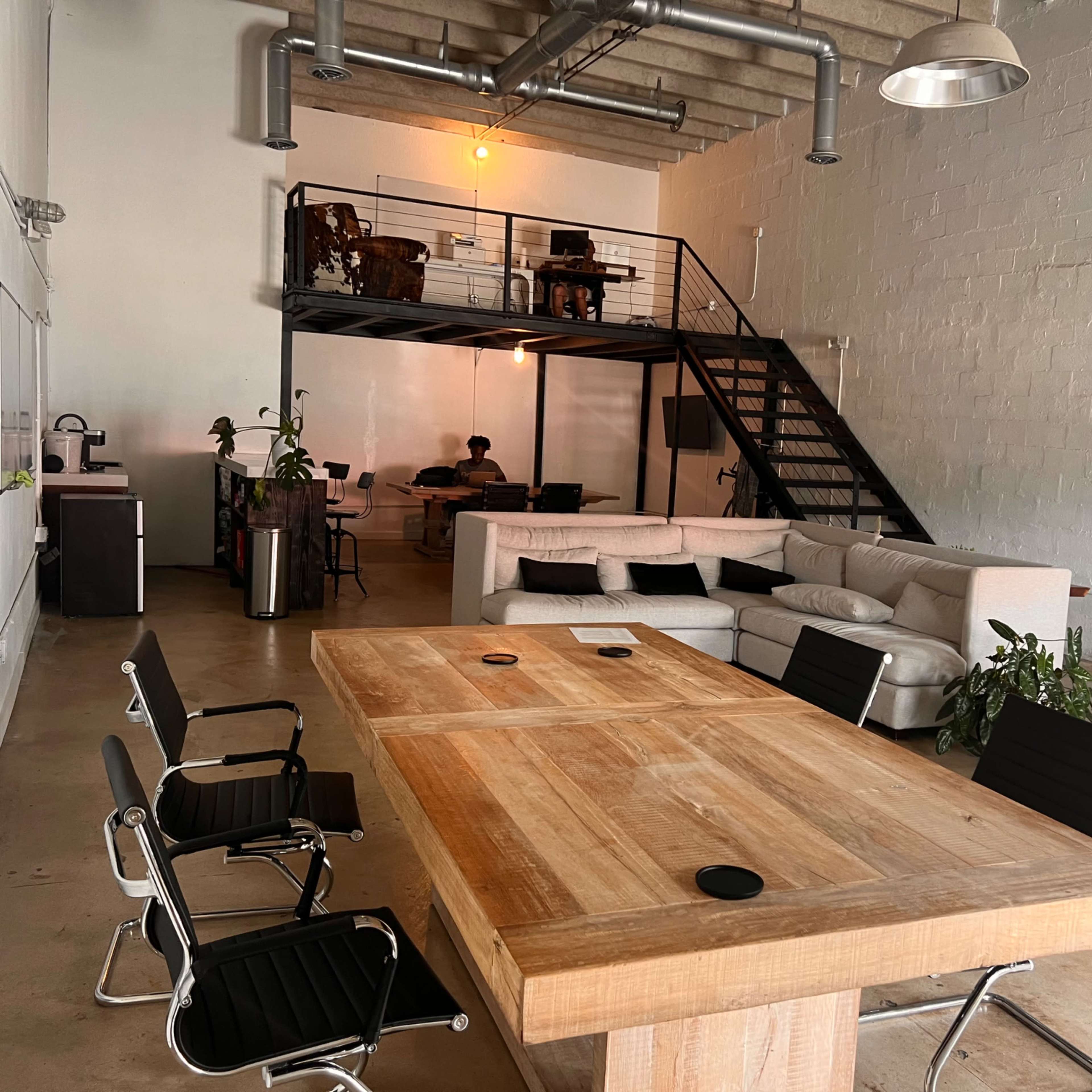 A spacious office features a wooden conference table surrounded by black chairs, with a lofted seating area and a couch in the background.