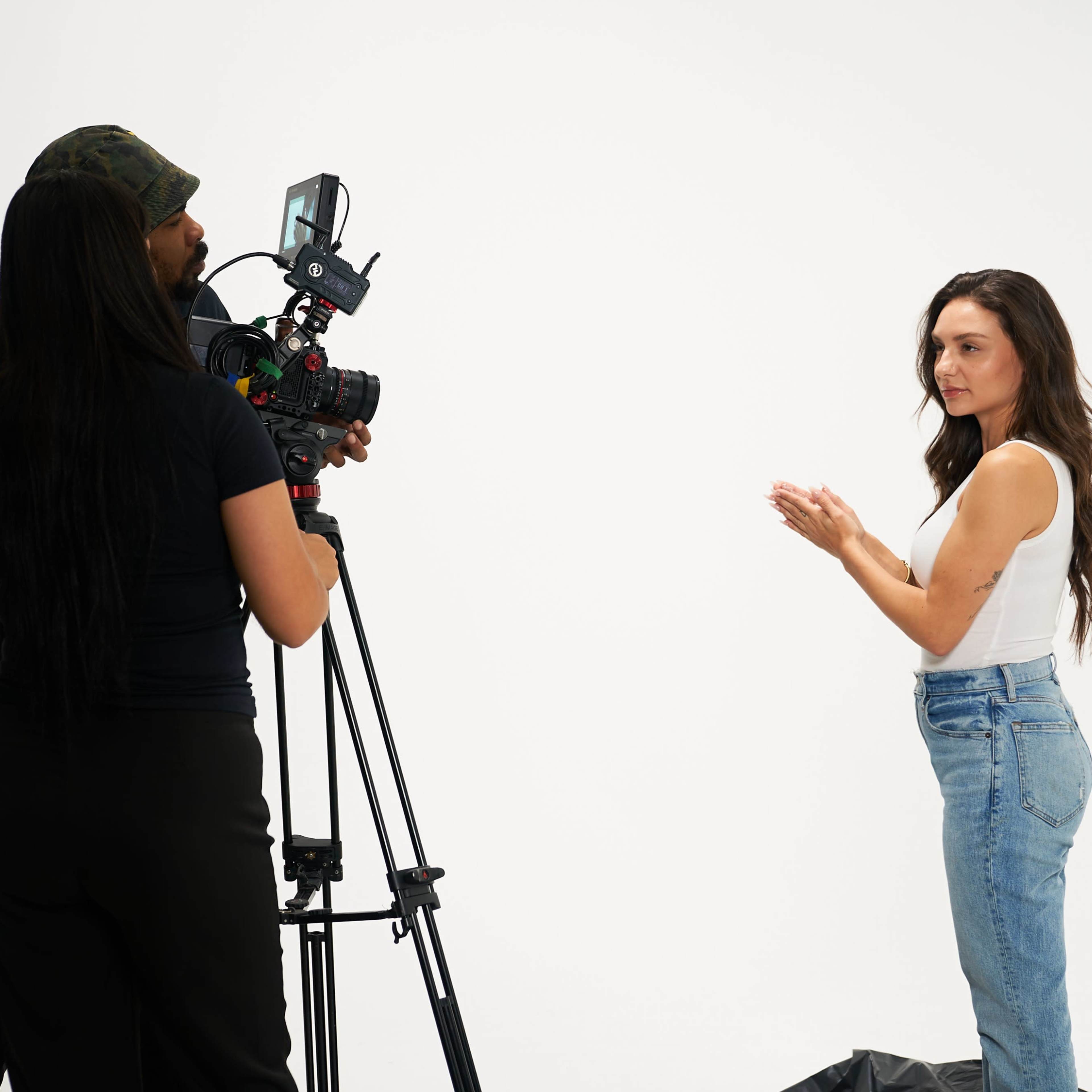 A woman stands in front of a camera while a crew member operates the equipment in a studio with a white backdrop.