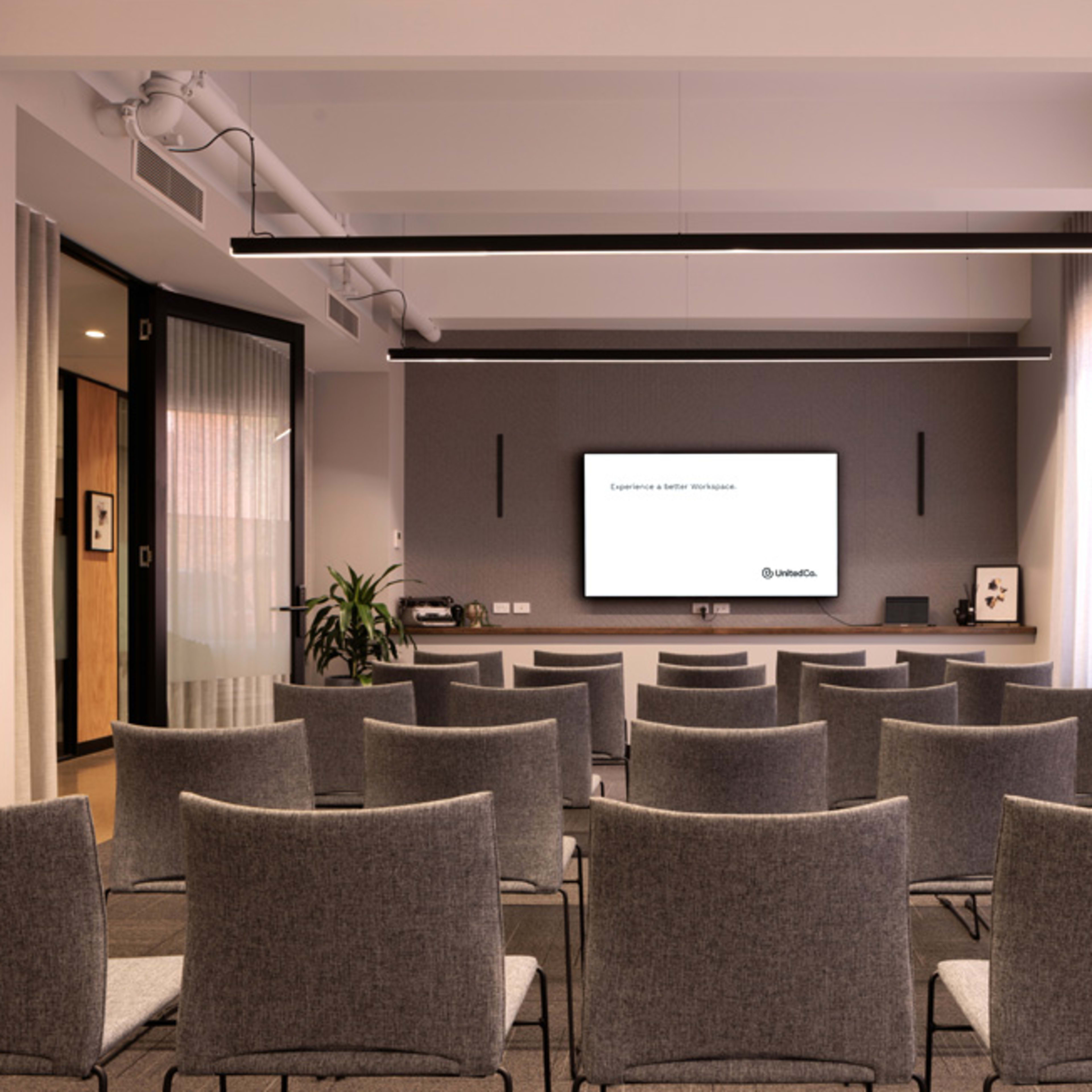 A modern conference room filled with gray chairs arranged in rows facing a mounted screen on a wall.