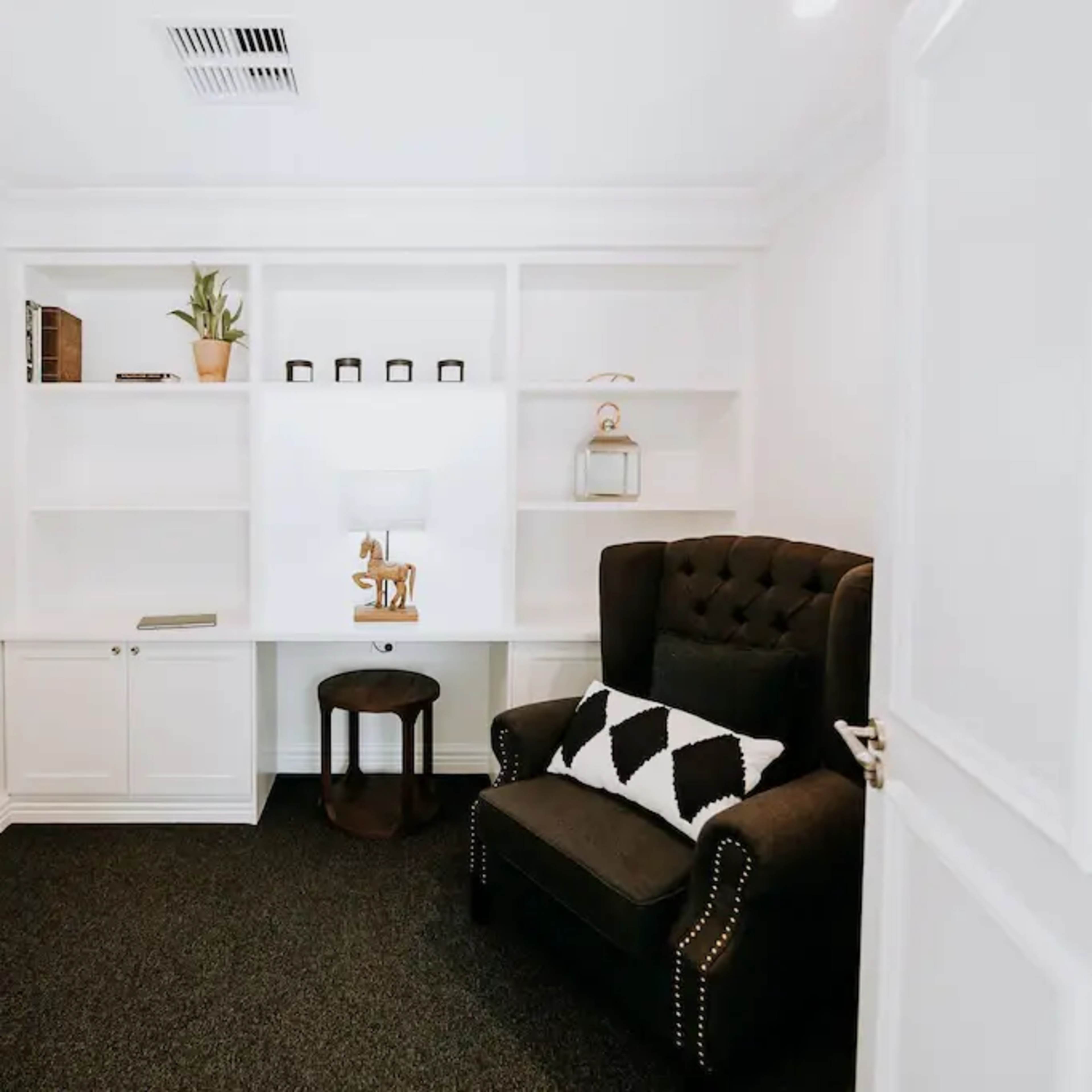 A small interior room with a black armchair, a round stool, a desk, and white shelves against the wall.