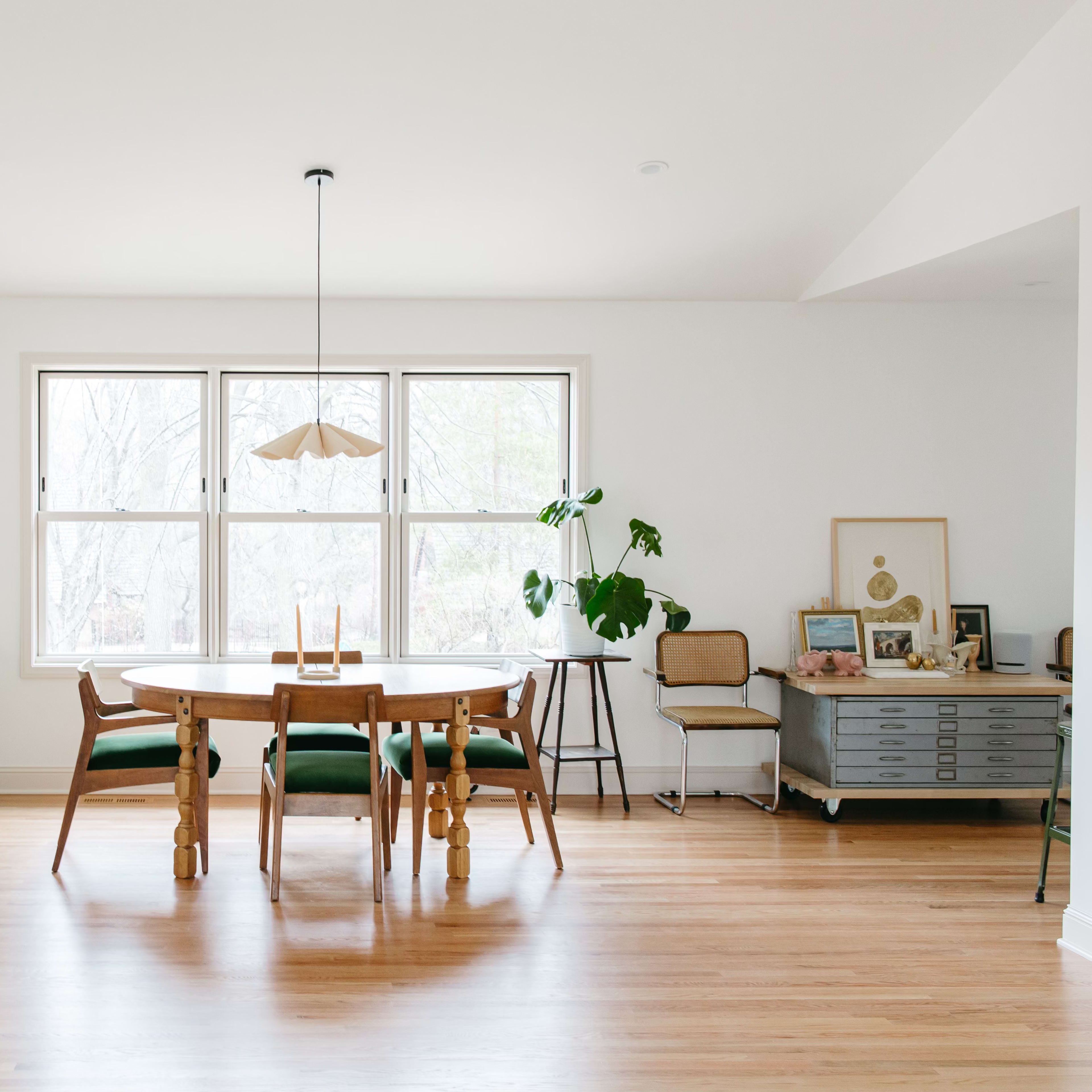 A modern dining area features a round table surrounded by wooden chairs, a large window providing natural light, and a mix of stylish furniture including a red accent chair and a sideboard.
