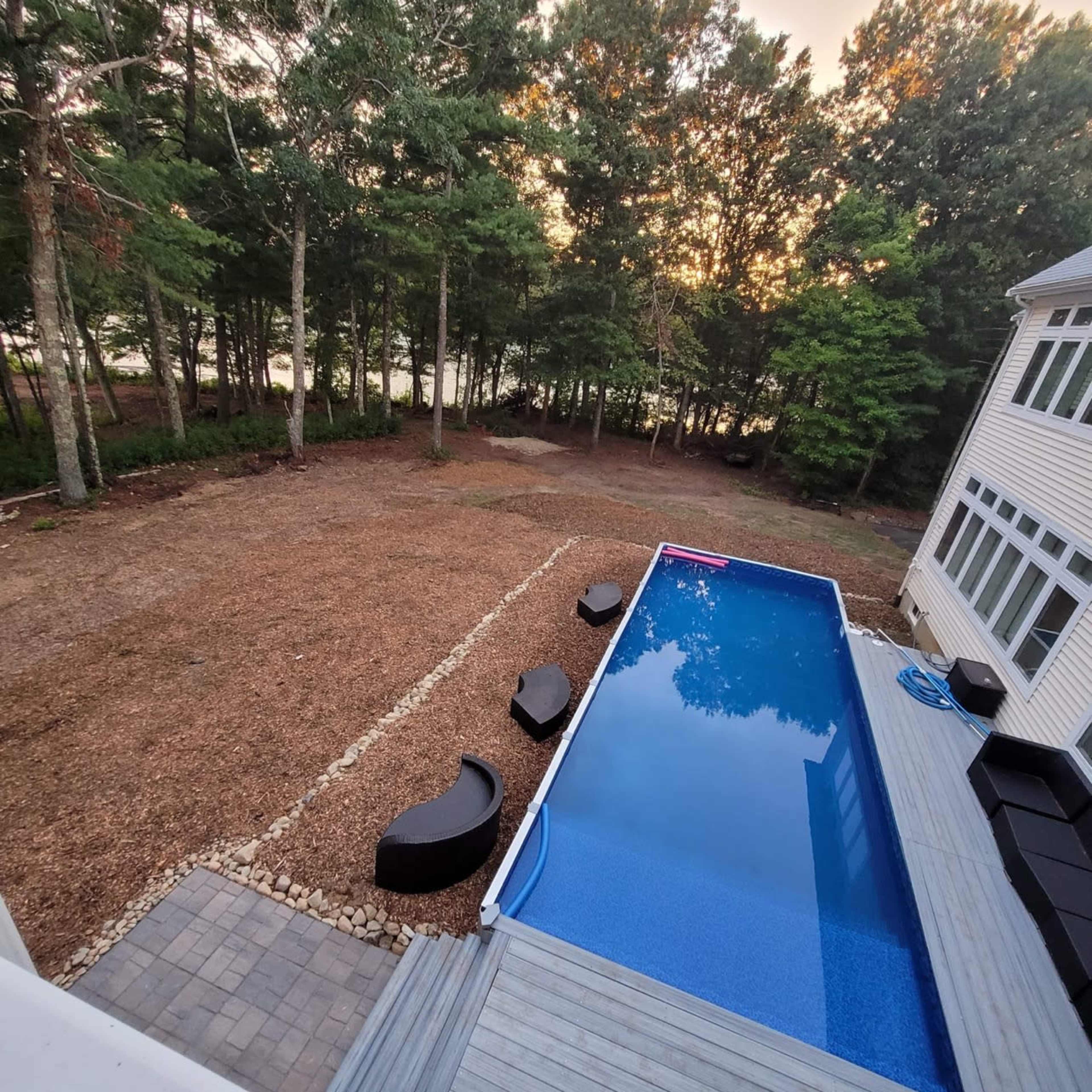 A modern rectangular swimming pool surrounded by a wooden deck and six curved lounge chairs in a wooded backyard.