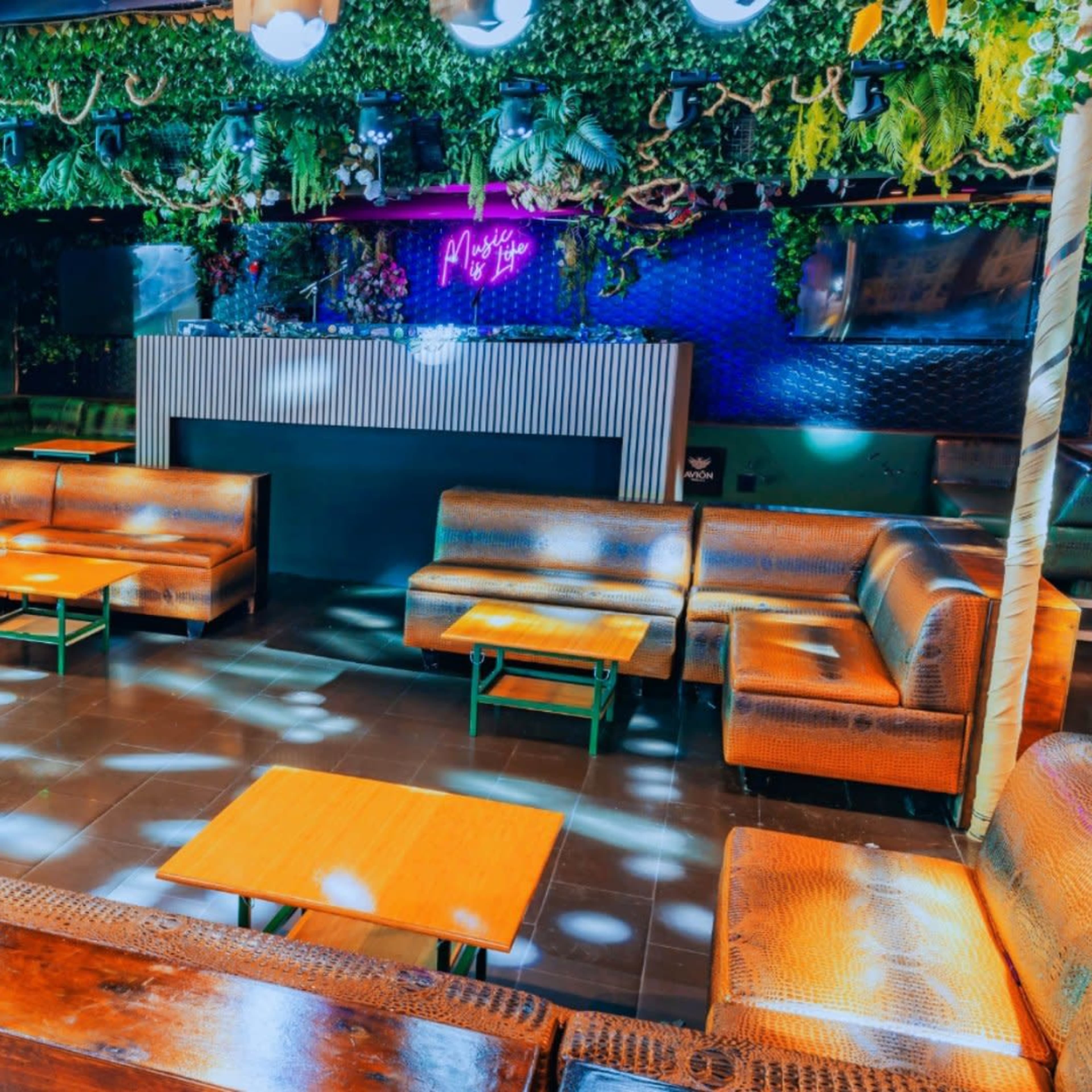 A vibrant lounge area with brown seating, wooden tables, and green foliage decorations under colorful lighting.
