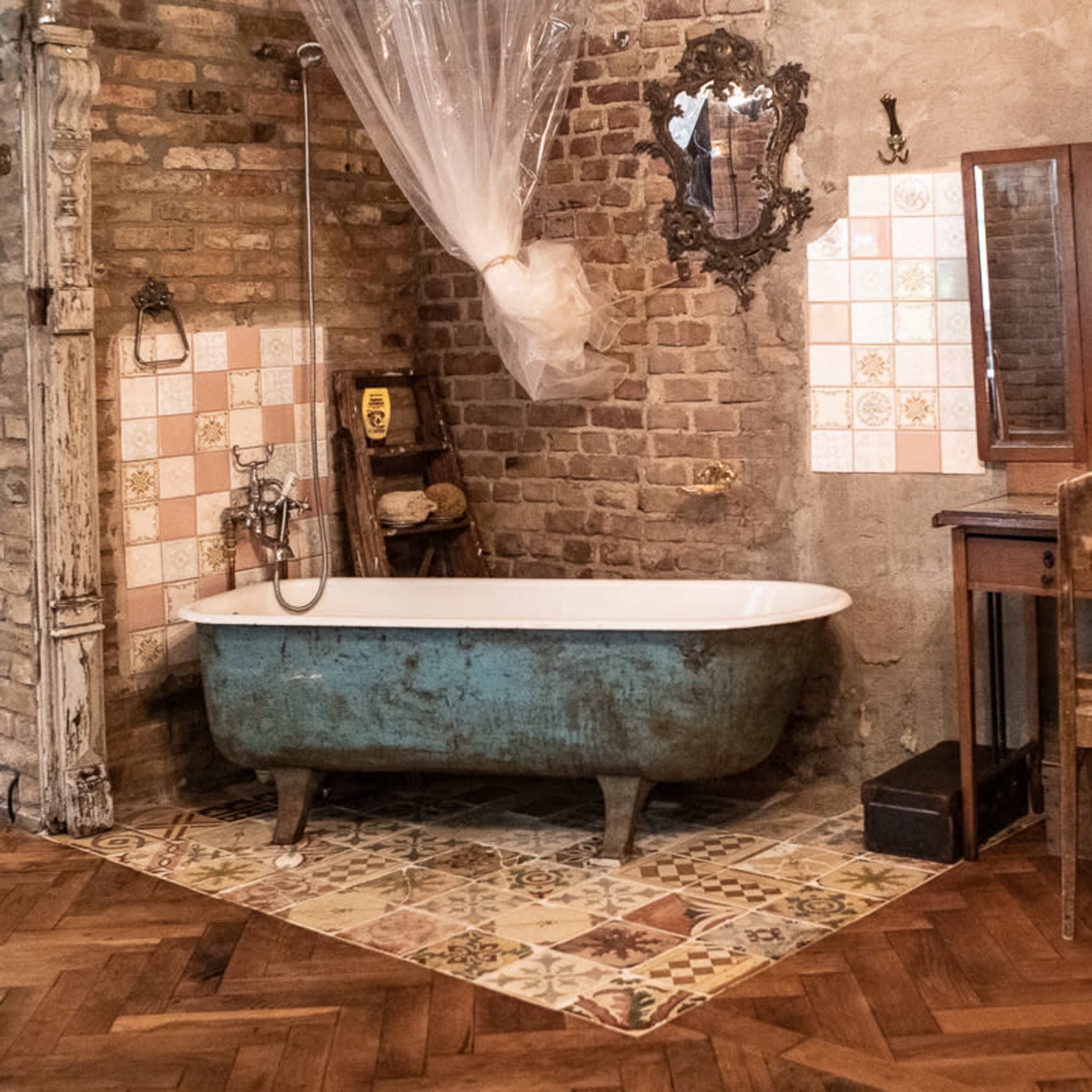 A vintage bathroom featuring a freestanding clawfoot bathtub, a wooden vanity with a mirror, and a wall with exposed brick and decorative tiles.
