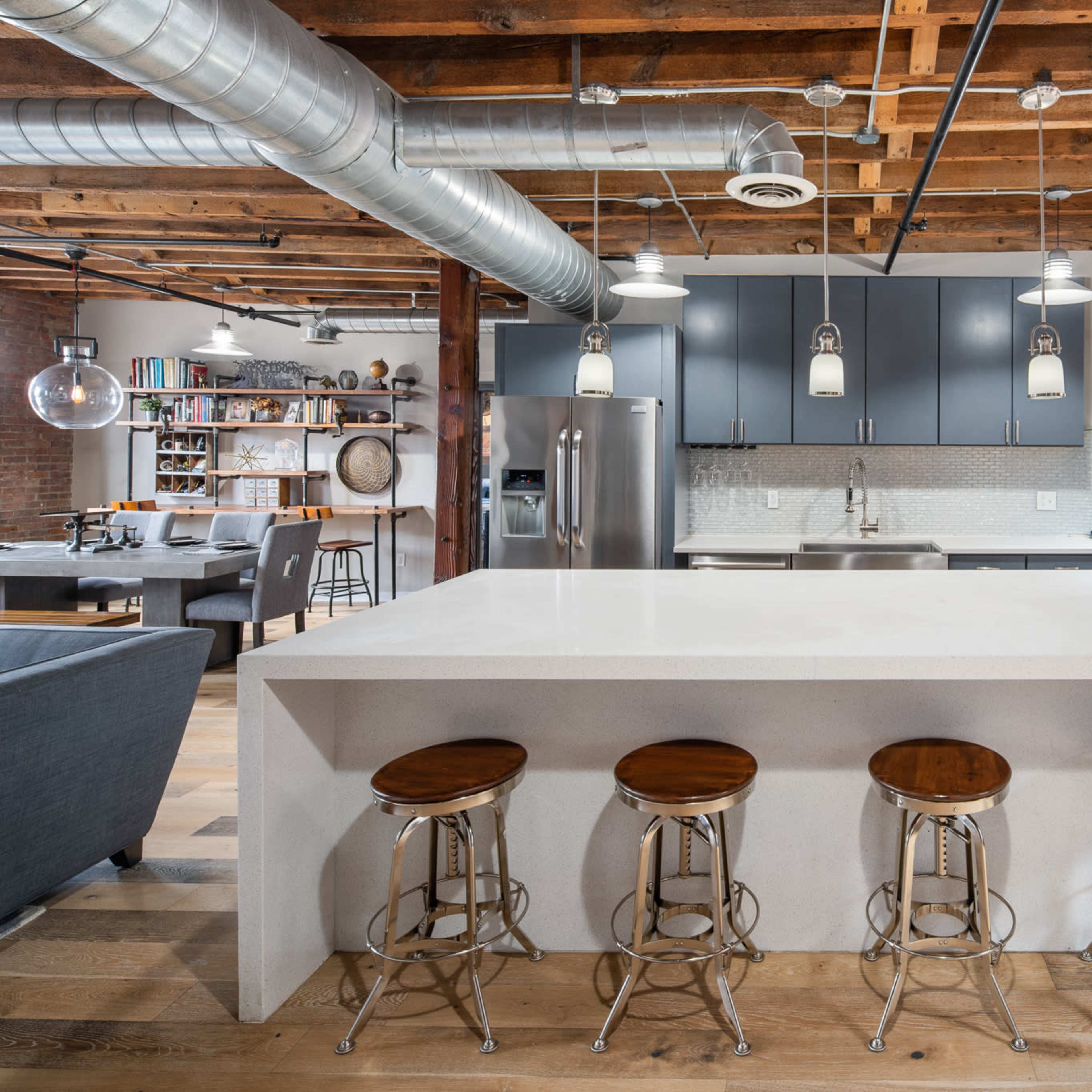 A modern kitchen with a central island, four metal and wooden stools, and exposed brick walls.