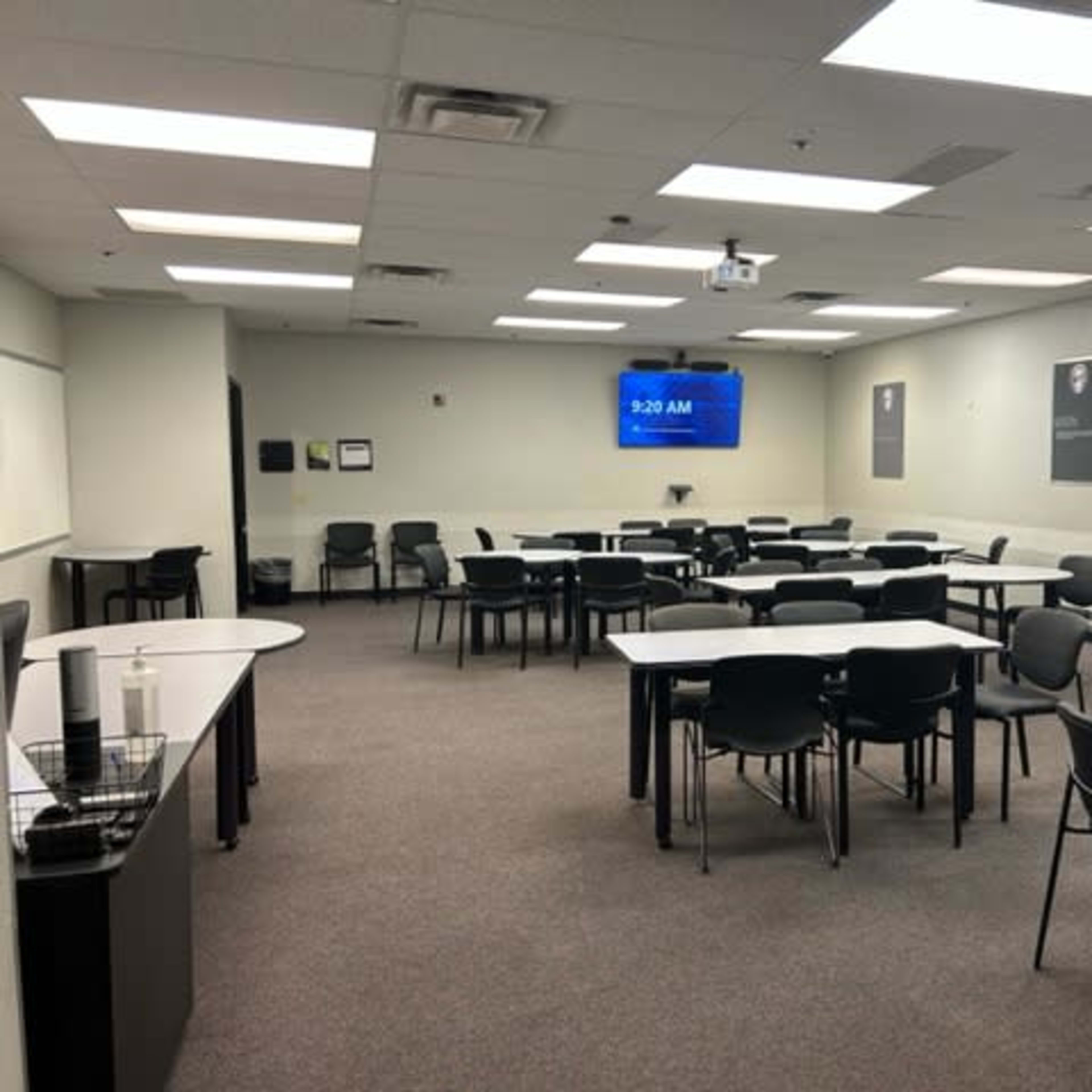 A spacious classroom is set up with multiple rows of tables and chairs, featuring a clock display showing 9:20 AM on a wall-mounted screen.