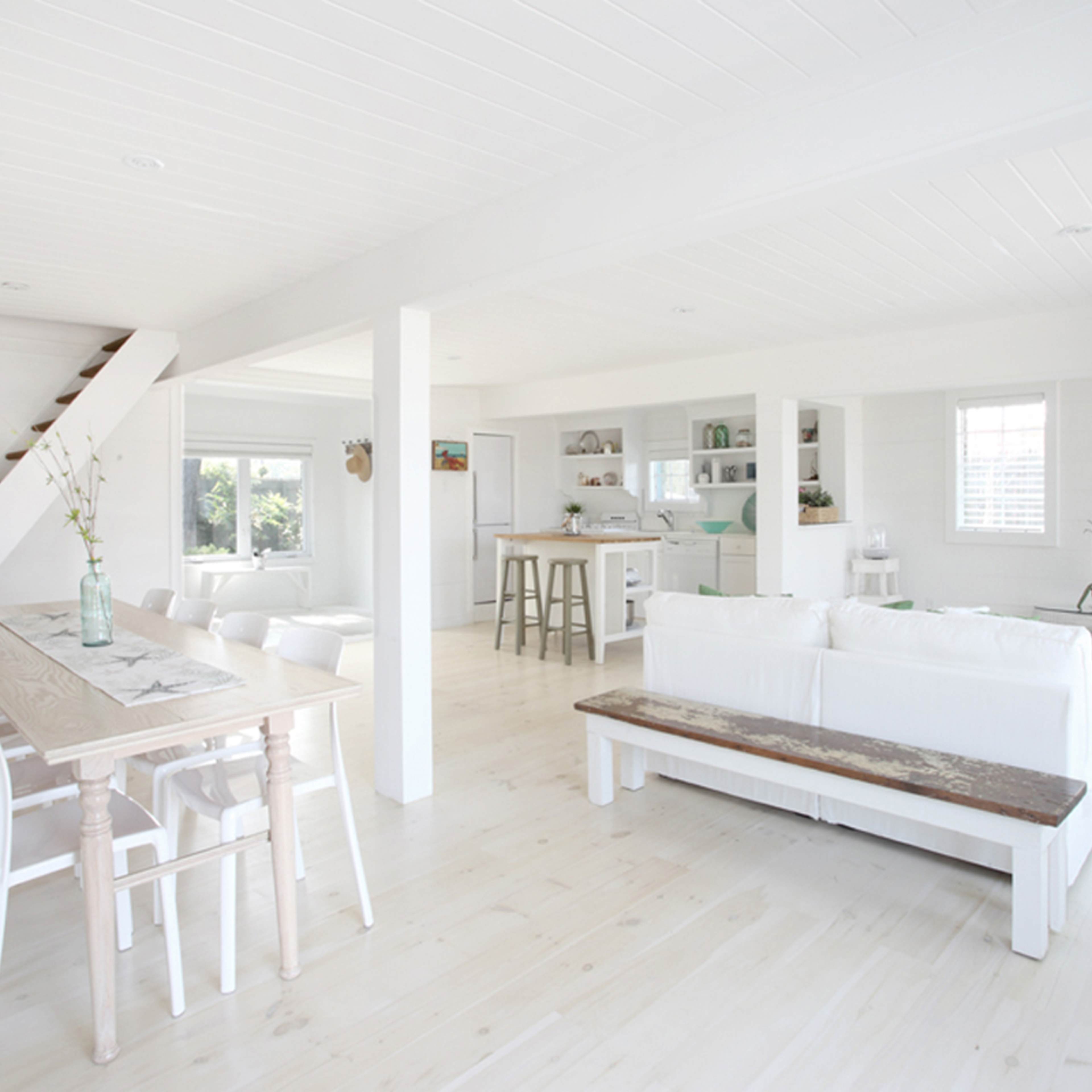 A bright, open living space with a white color scheme, featuring a dining area with a wooden table and chairs, a sitting area with a white sofa, and a staircase in the background.