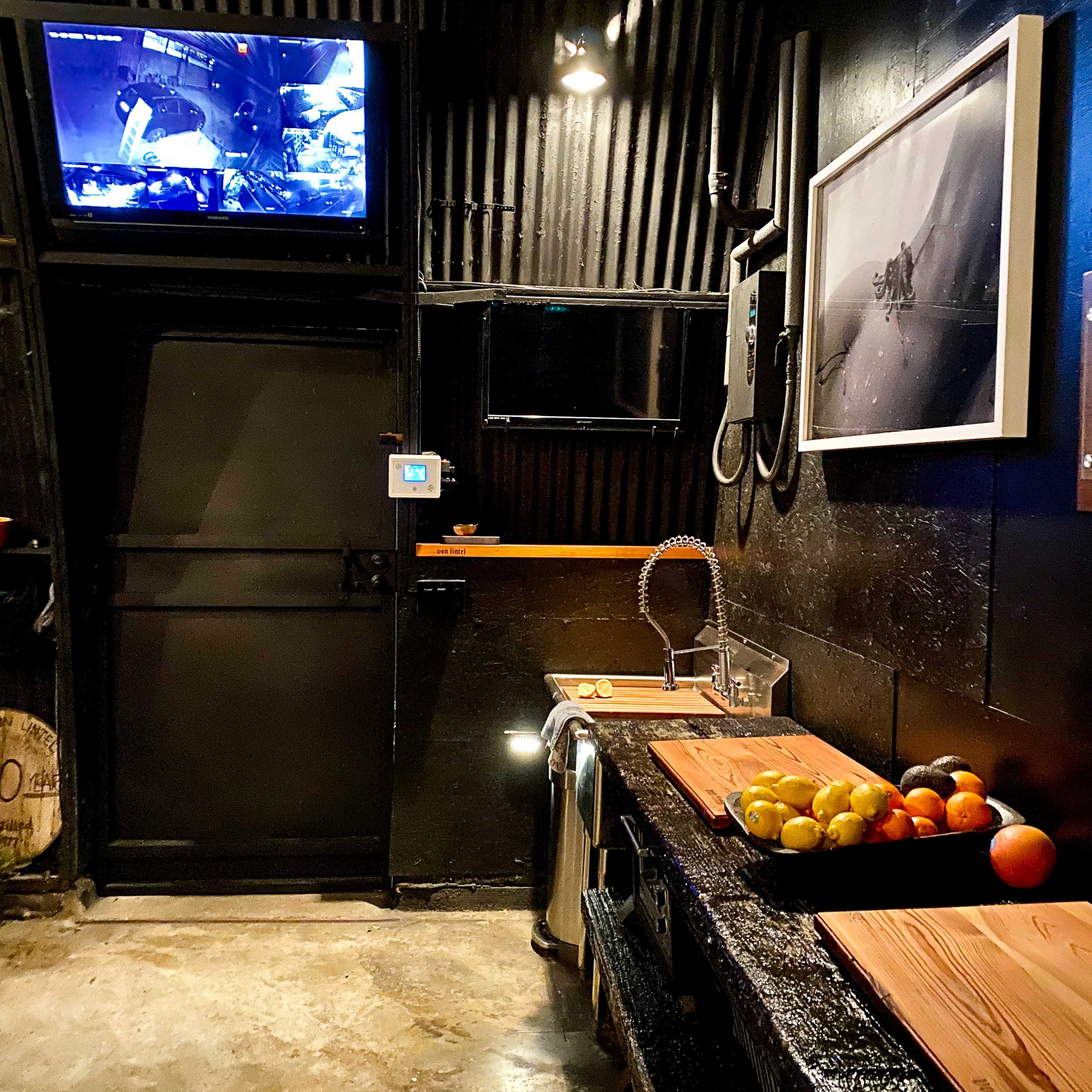 A dark kitchen with a stainless steel sink, wooden countertops, a bowl of colorful fruits, and two wall-mounted screens displaying video feeds.