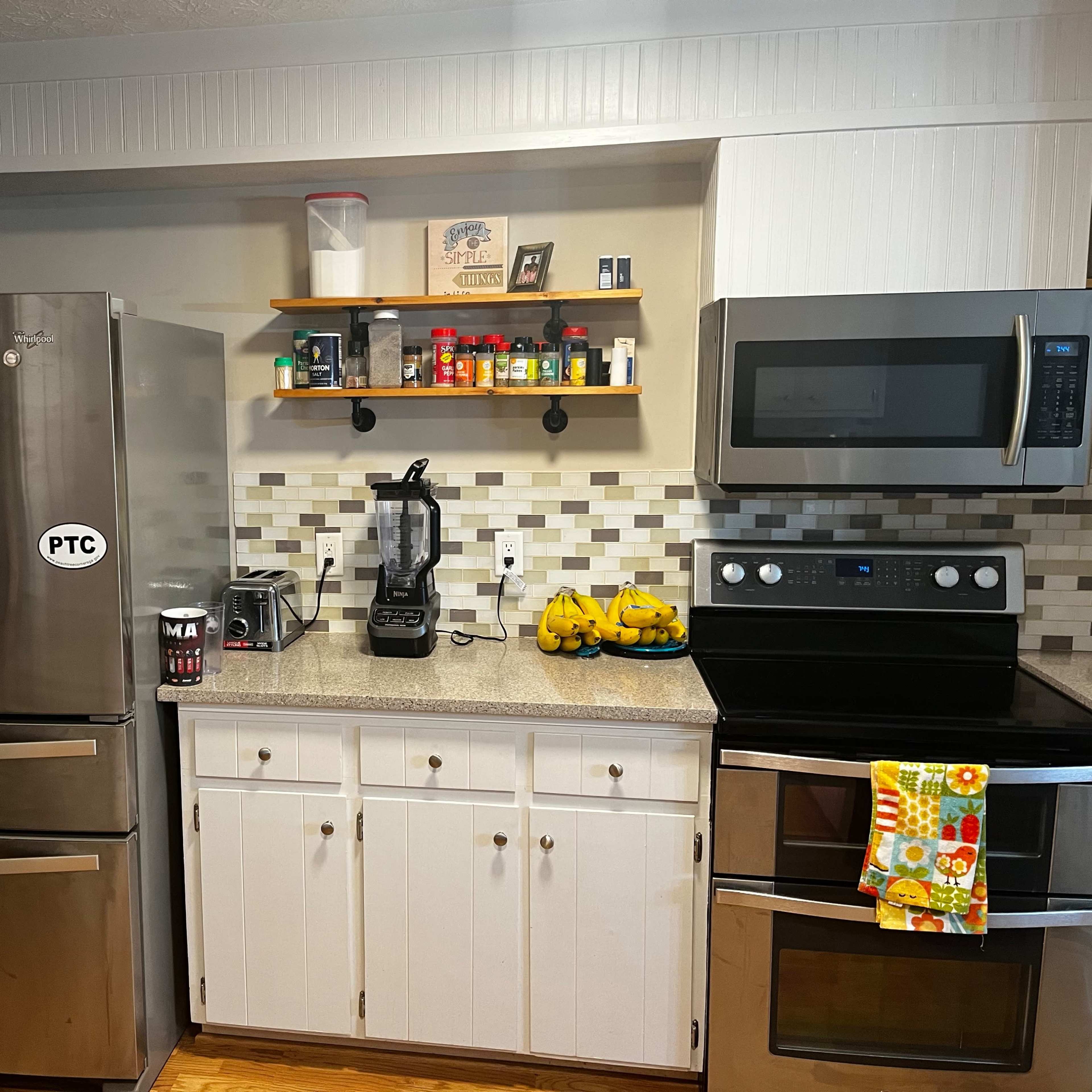 A kitchen features stainless steel appliances, a countertop with a blender and a bowl of bananas, and shelves holding various spices and condiments.