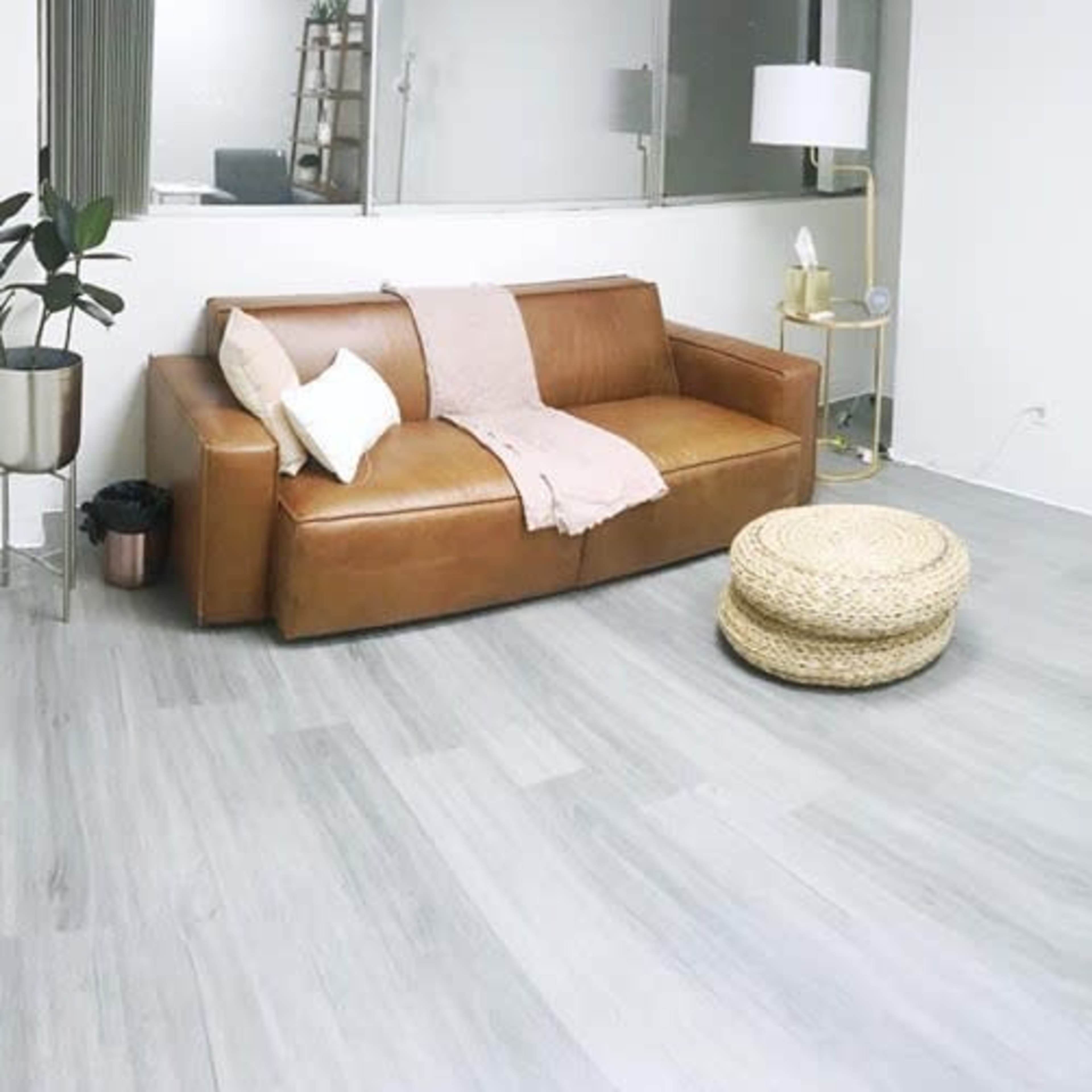 A modern, minimalist living room with a brown leather sofa, a round woven pouf, a gray armchair, and a white coffee table on light hardwood flooring.