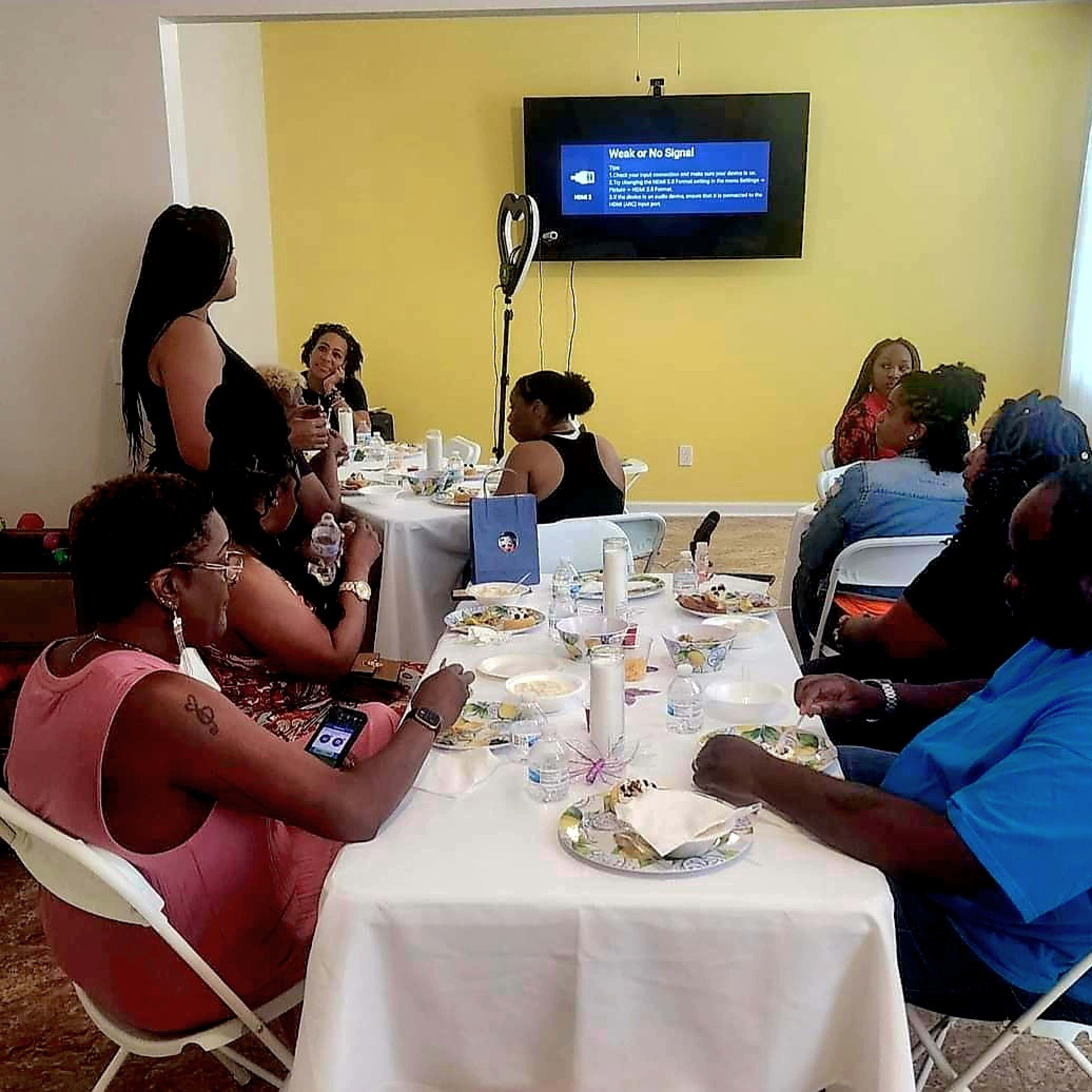 A group of people sits around tables sharing a meal while a woman stands and addresses the audience in a brightly lit room.