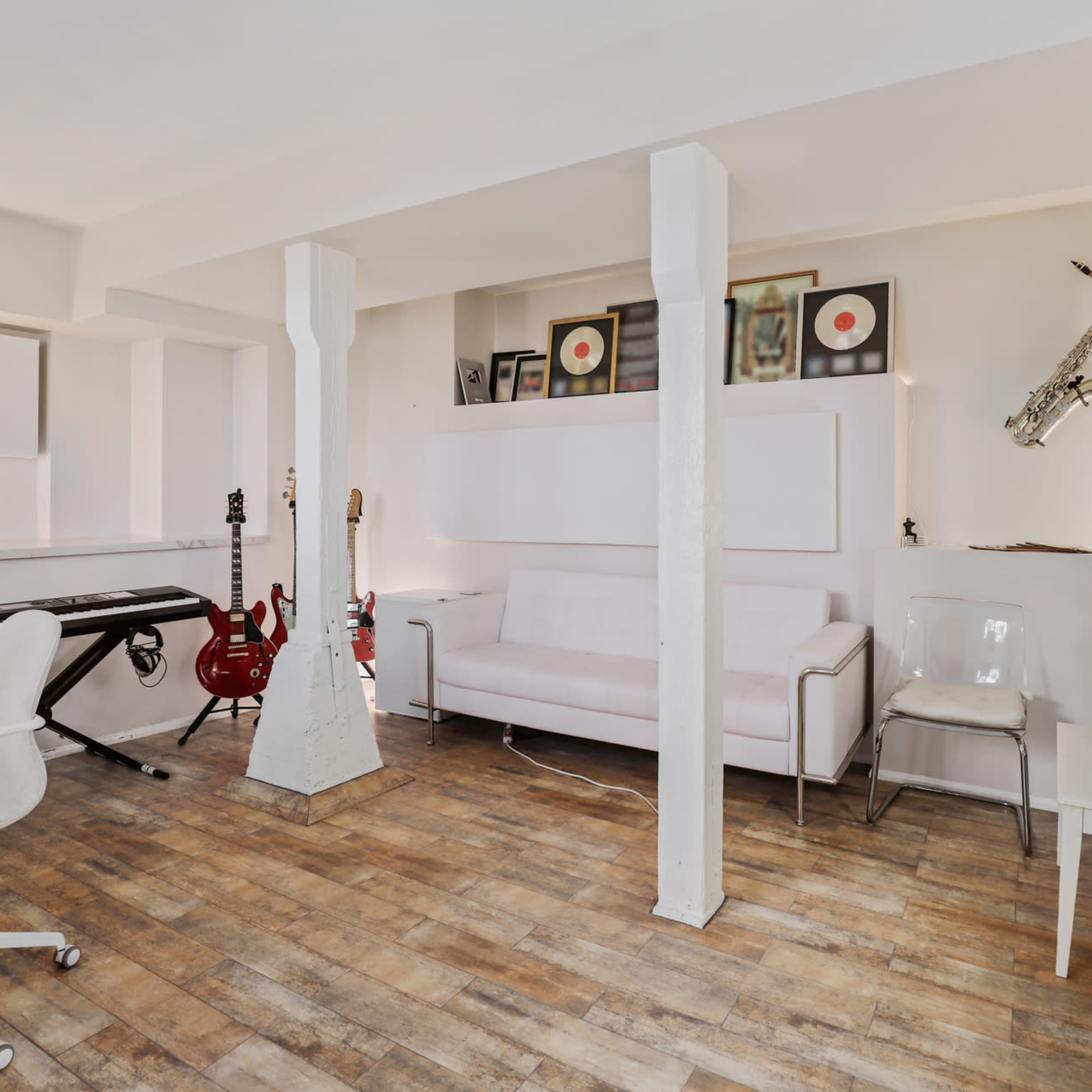 A modern home studio features a keyboard, electric guitars, a white couch, and a saxophone mounted on the wall, all arranged in a bright setting.