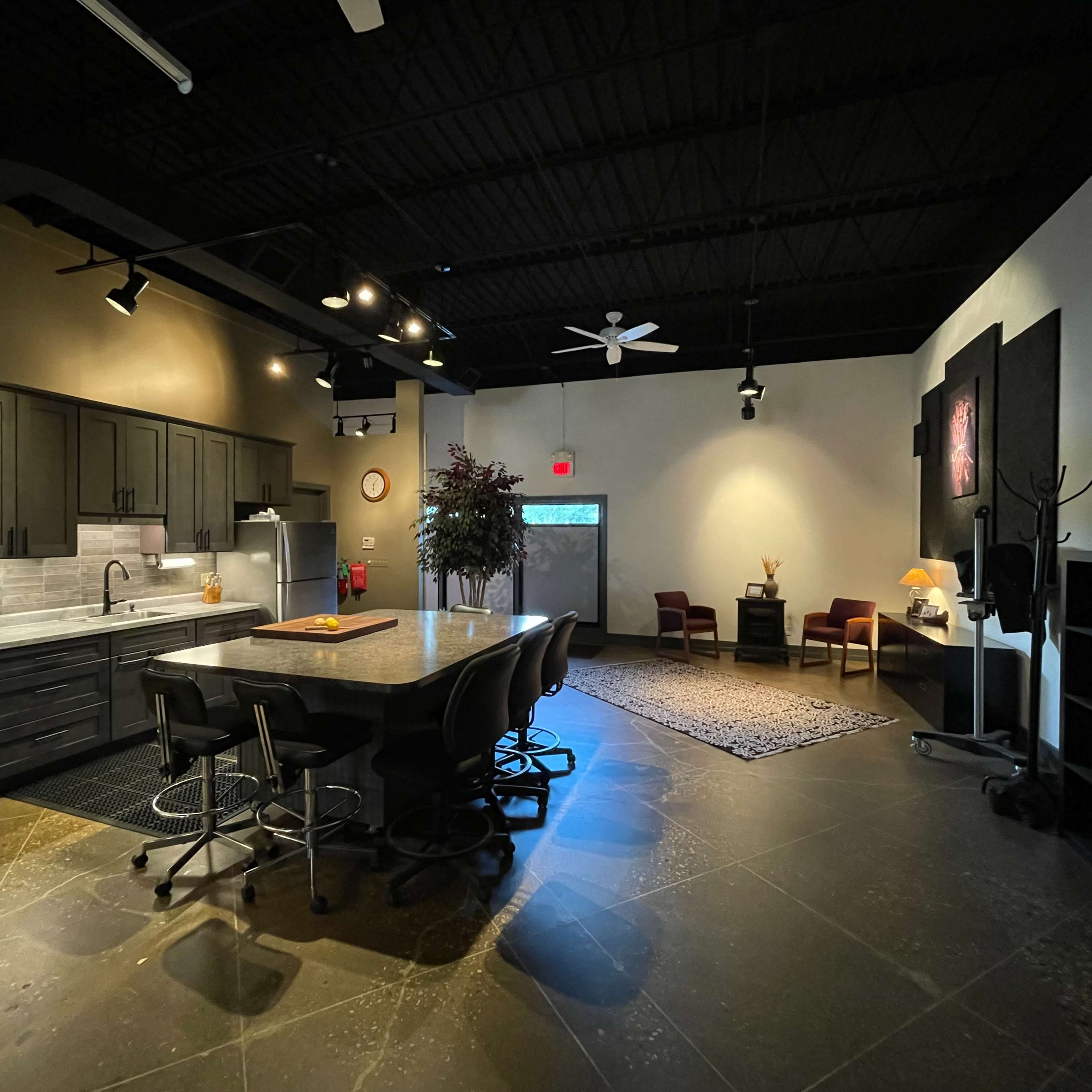 A modern kitchen with dark cabinetry, stainless steel appliances, and a large island, adjacent to a spacious living area with two chairs and a rug.