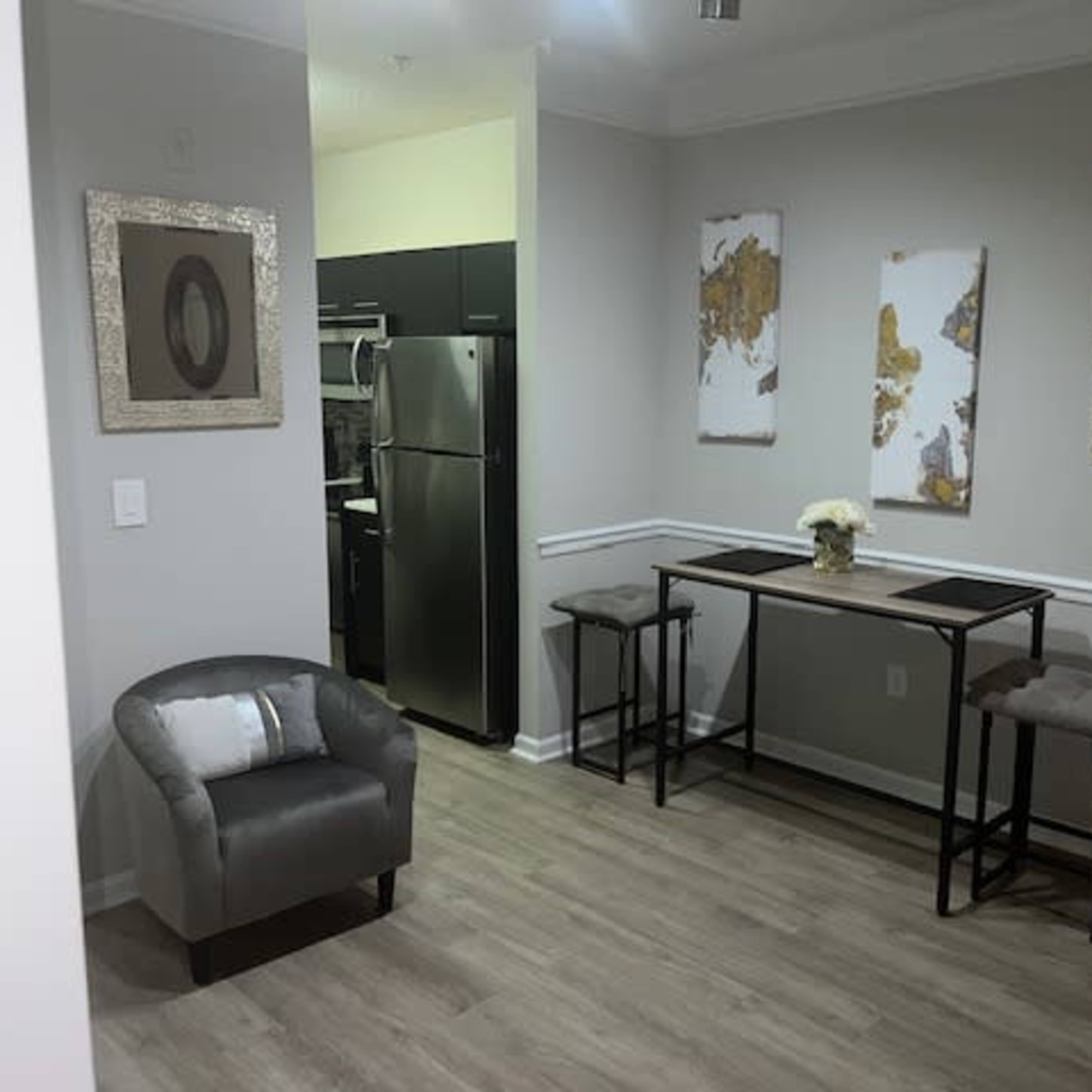A dining area featuring a small table with two stools, a chair in the corner, and three framed wall decorations, along with a kitchenette visible in the background.