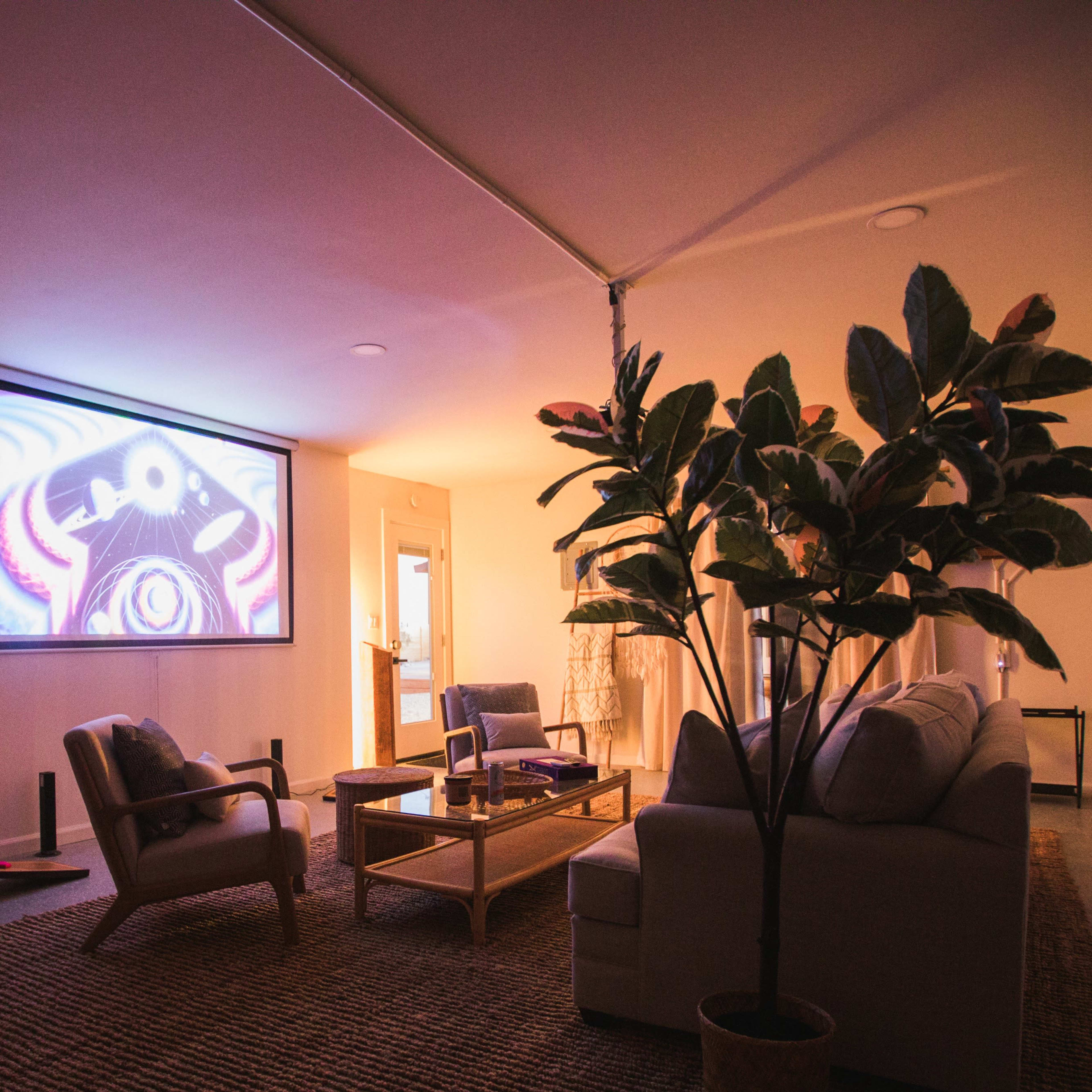 A living room features a projector screen displaying colorful artwork, with a sofa, armchairs, and a large potted plant arranged around a rectangular coffee table.