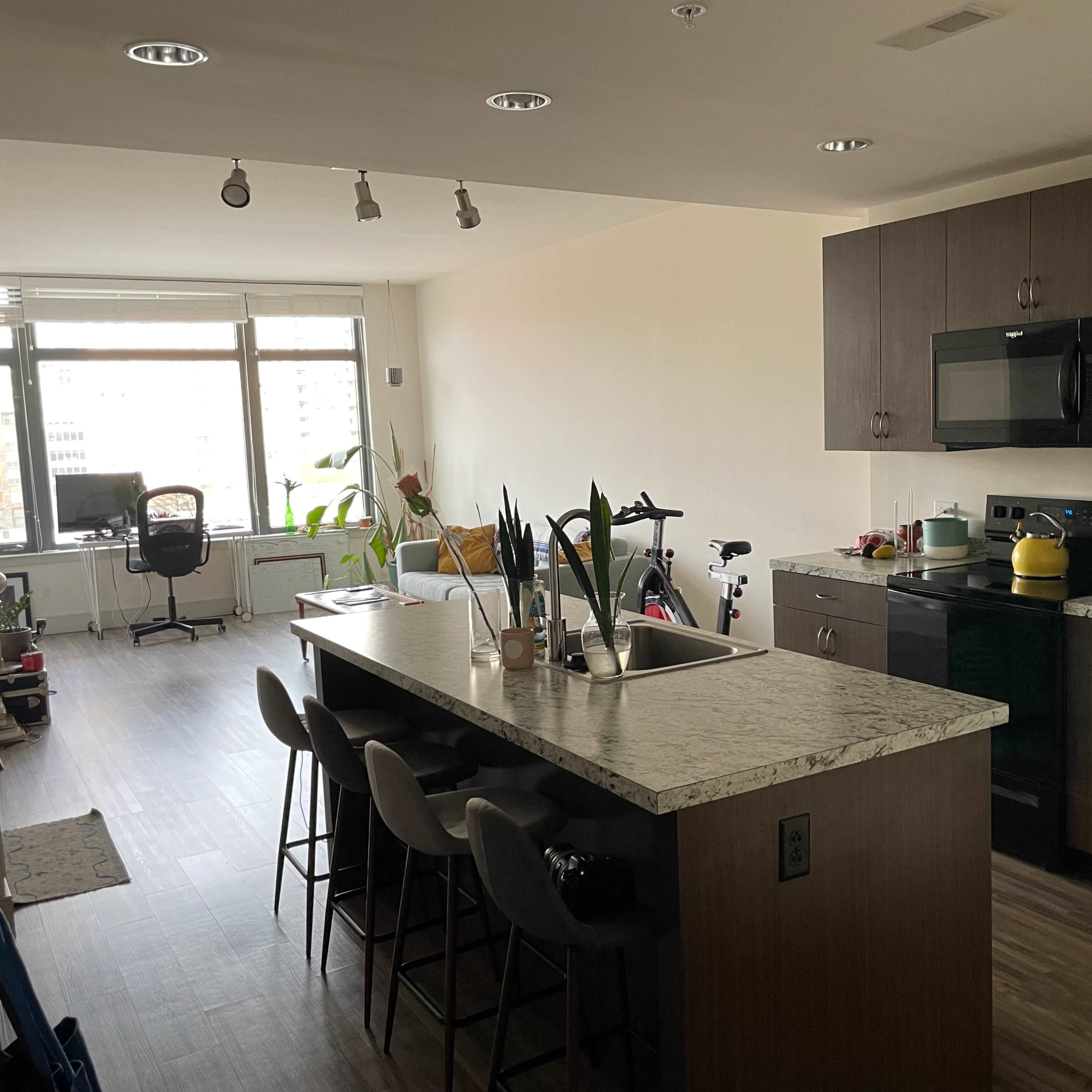 A modern kitchen with an island, four bar stools, and an adjacent living area featuring a desk and plants by the window.