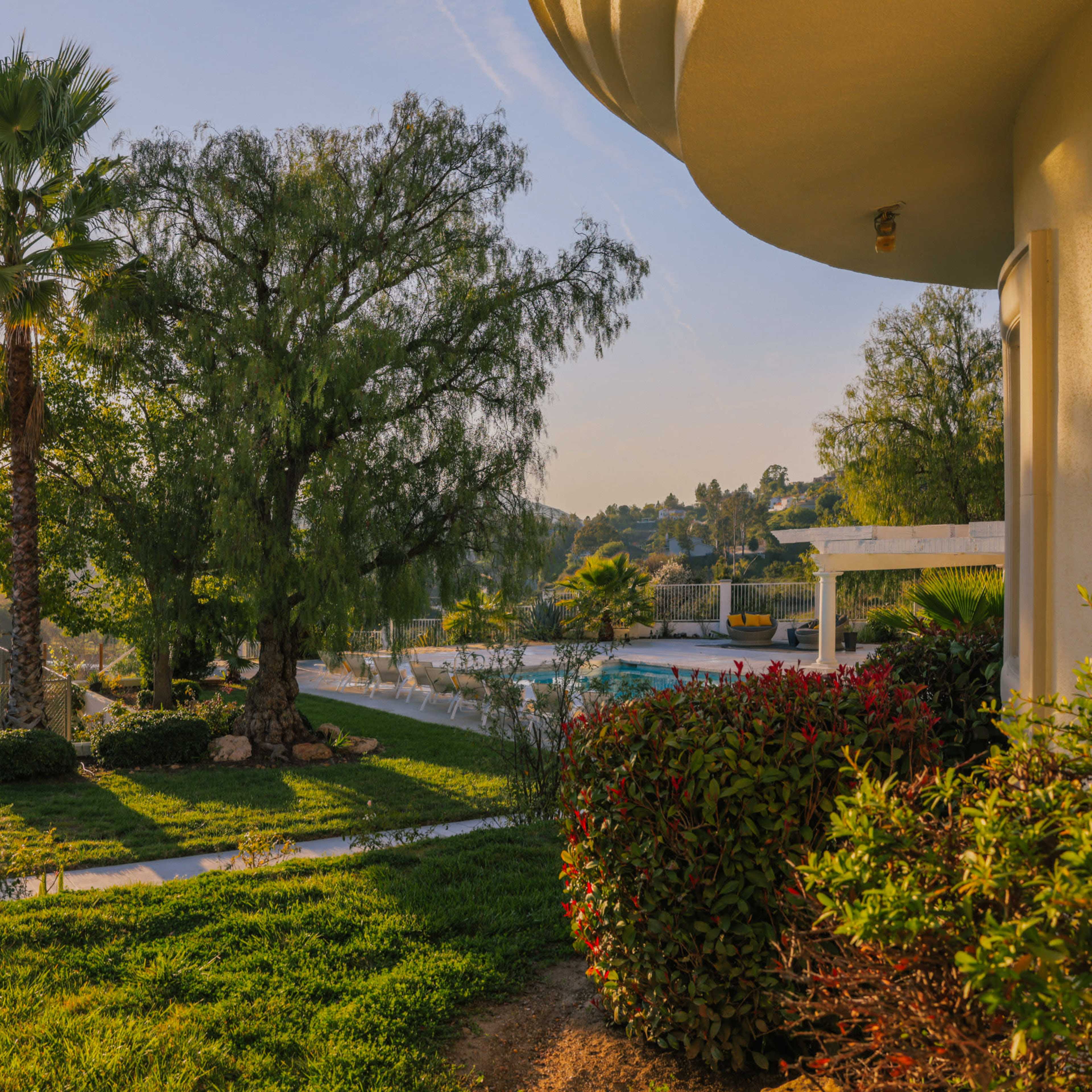 The scene features a landscaped garden with palm trees and a swimming pool surrounded by hills and greenery under a clear sky.