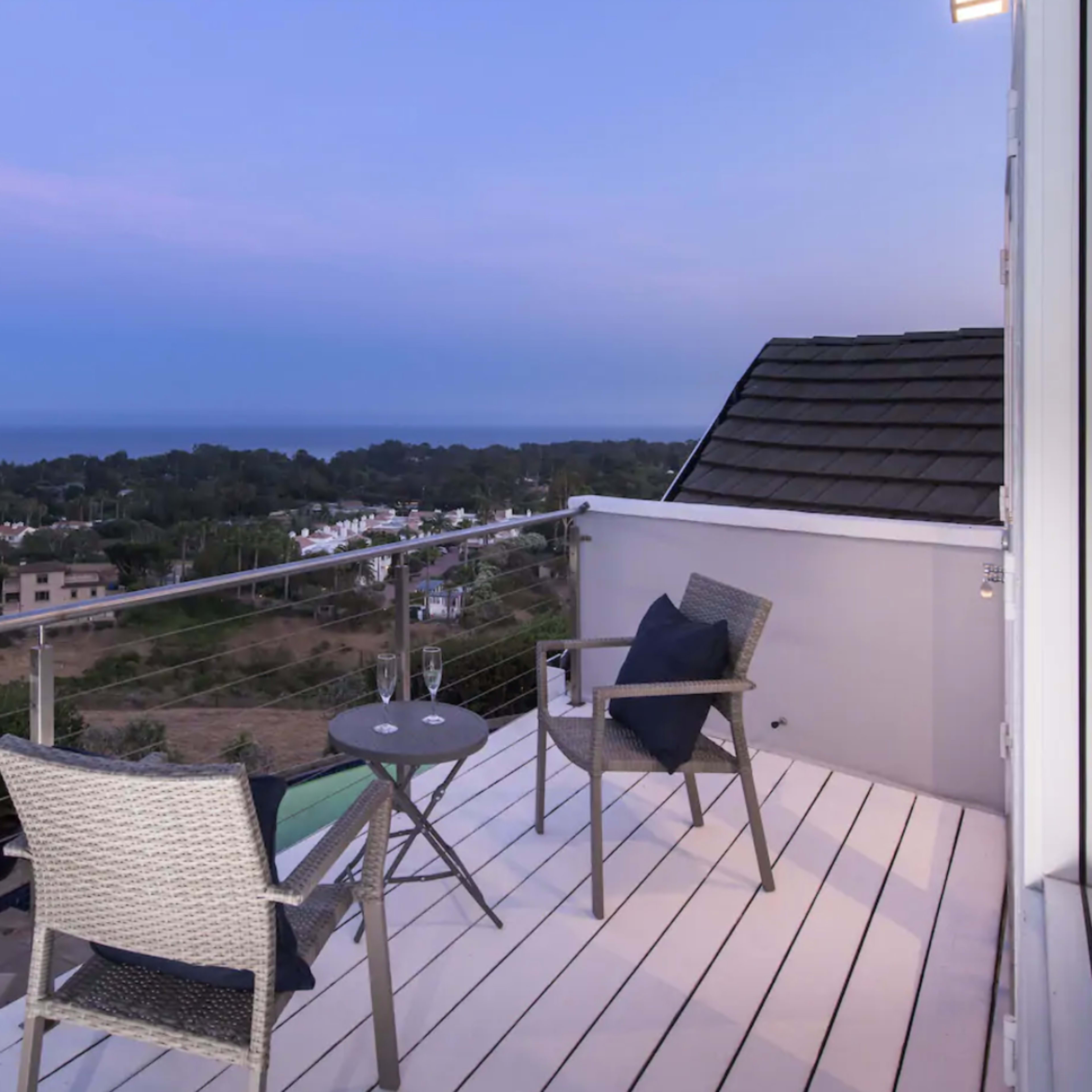 A balcony features two chairs and a small table, overlooking a scenic view of a coastal area at dusk.