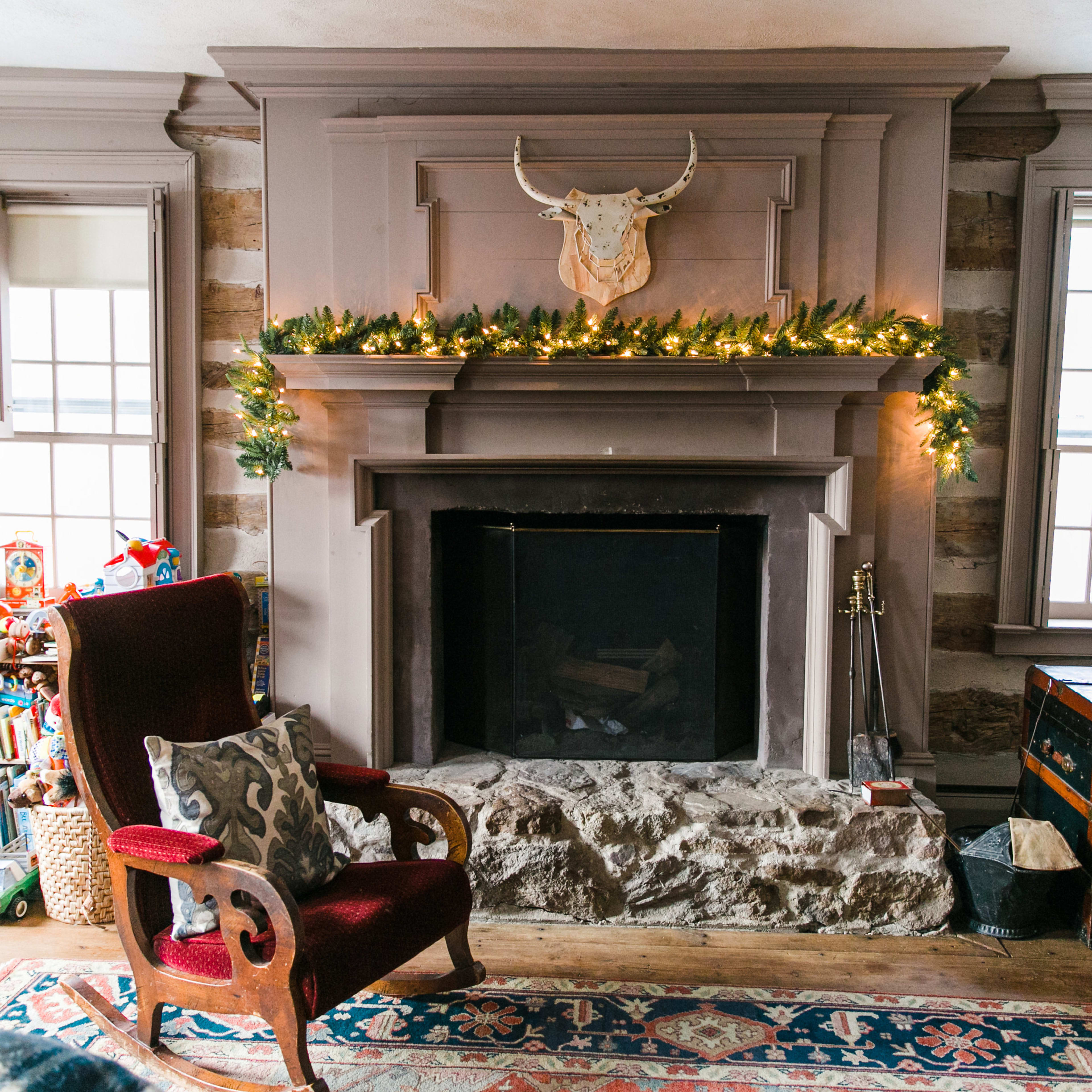 The living room features a stone fireplace adorned with greenery and lights, surrounded by logs on the wall and various toys scattered on the floor.