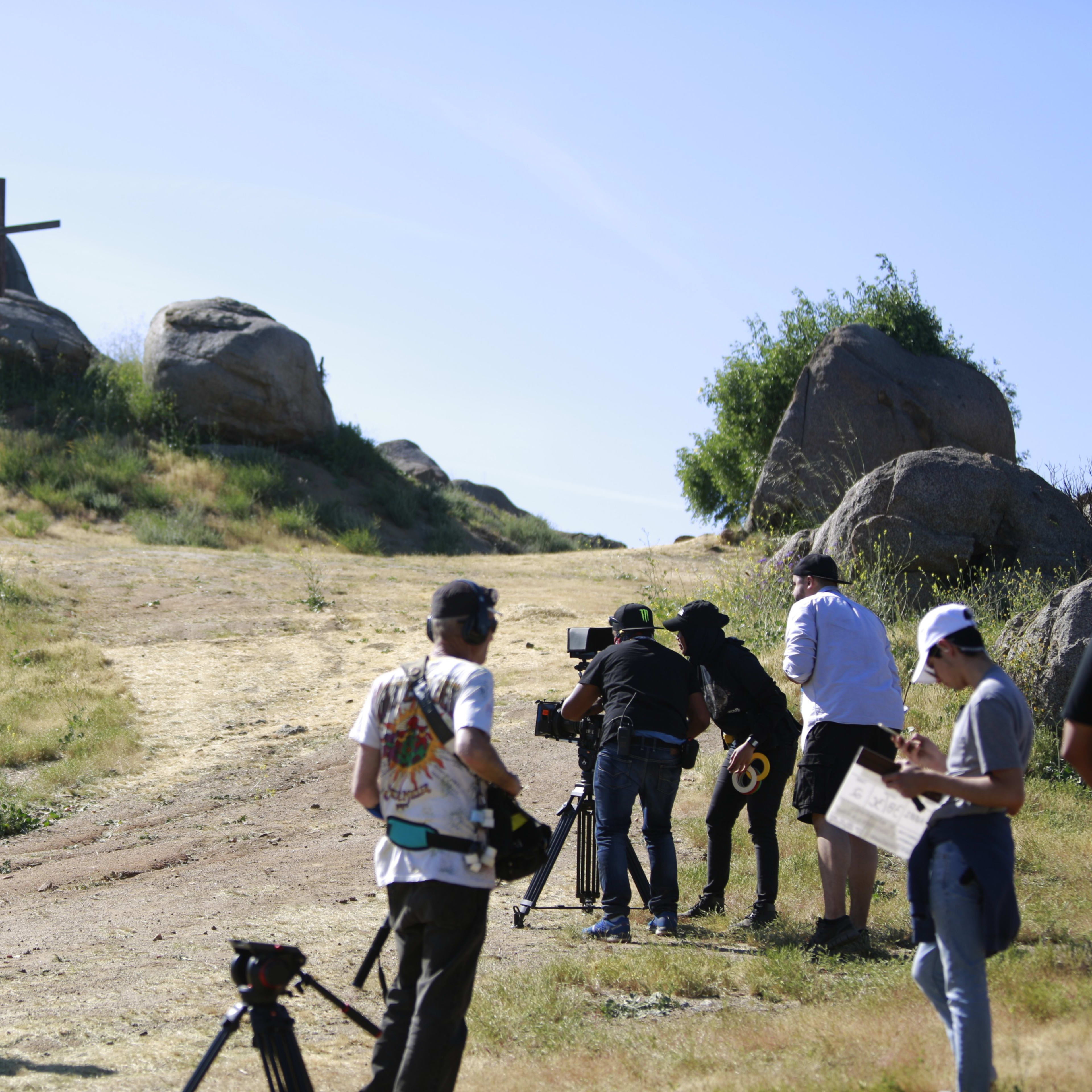 A film crew prepares to shoot a scene near three crosses on rocky terrain under a clear blue sky.