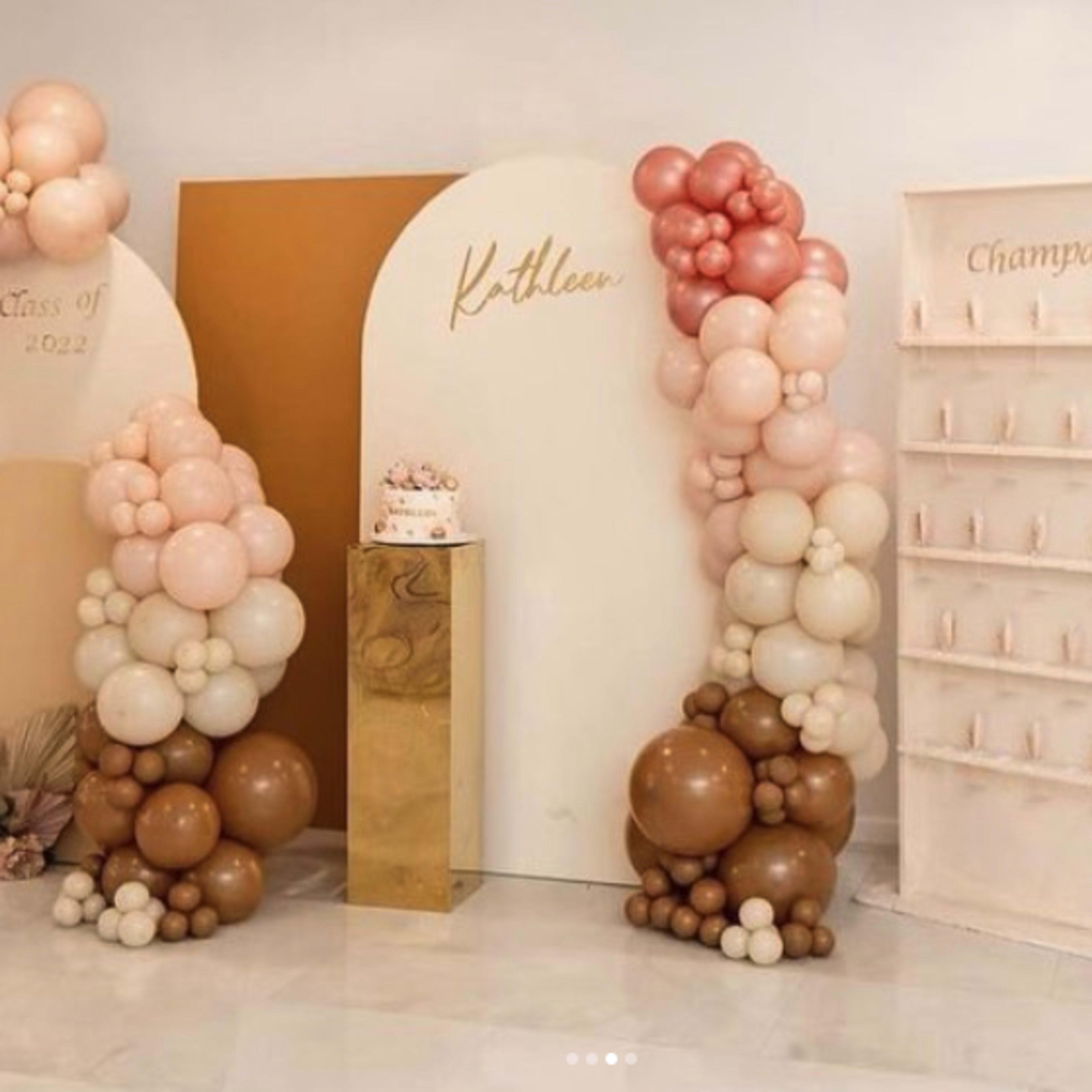 The scene features a festive decor setup with balloon arches, a gold pedestal displaying a cake, and a champagne wall, all arranged for a graduation celebration.