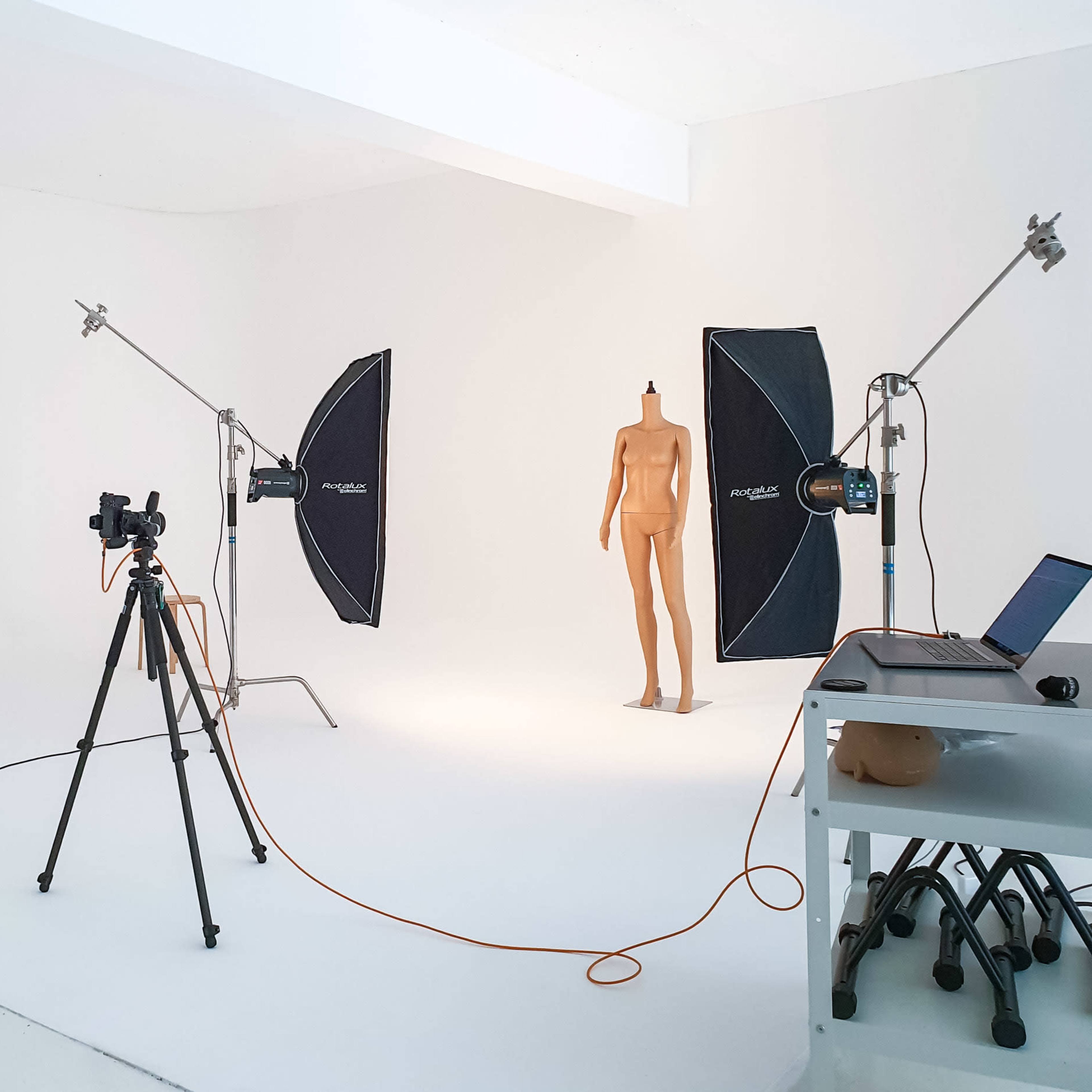 A photography studio with a beige mannequin, two softbox lights, a camera on a tripod, and a laptop on a cart.