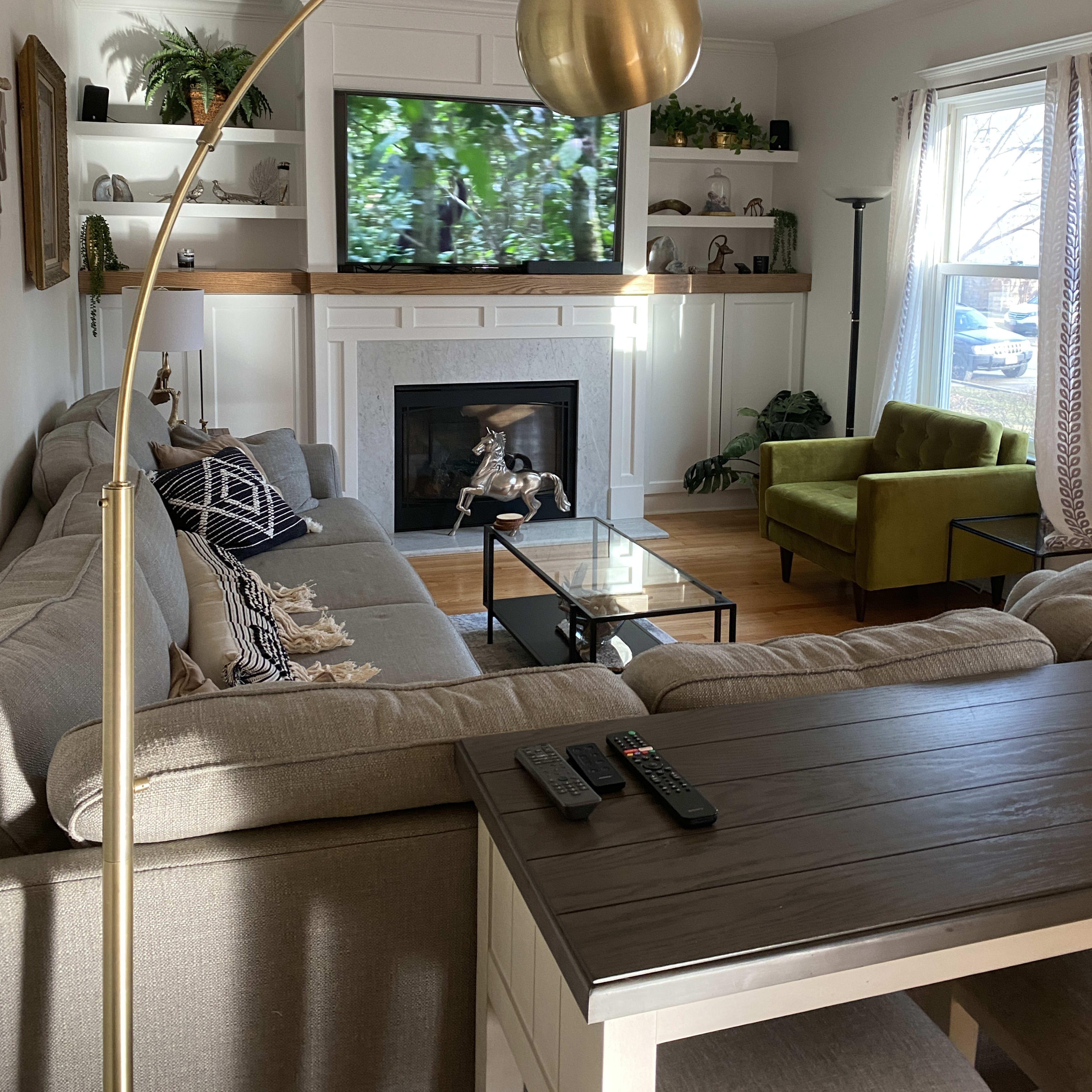 The living room features a sectional sofa, a coffee table, and a large television mounted above a fireplace, with greenery visible through the window.