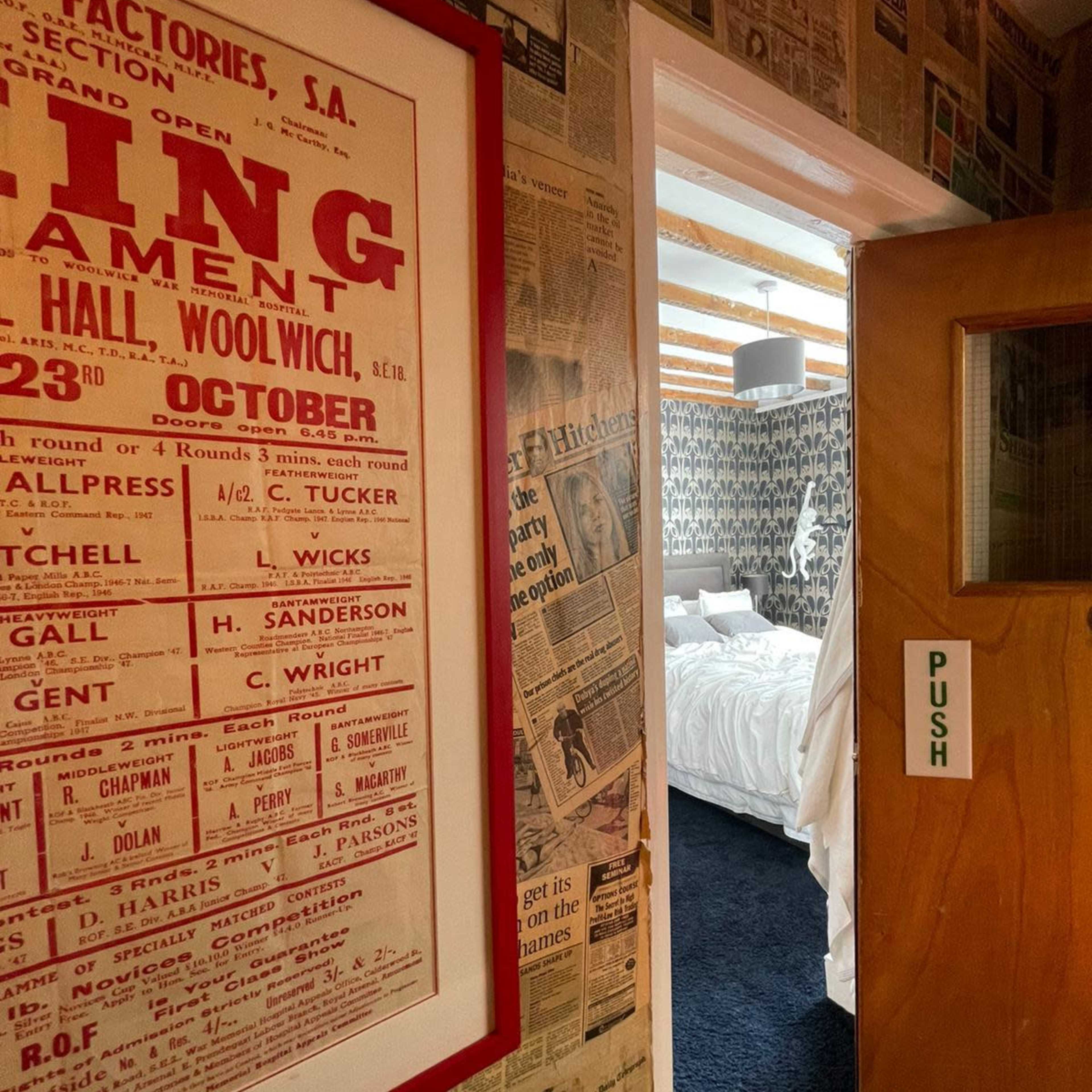 A hallway with a wall covered in newspaper clippings, leading to a room with a bed and patterned wallpaper.