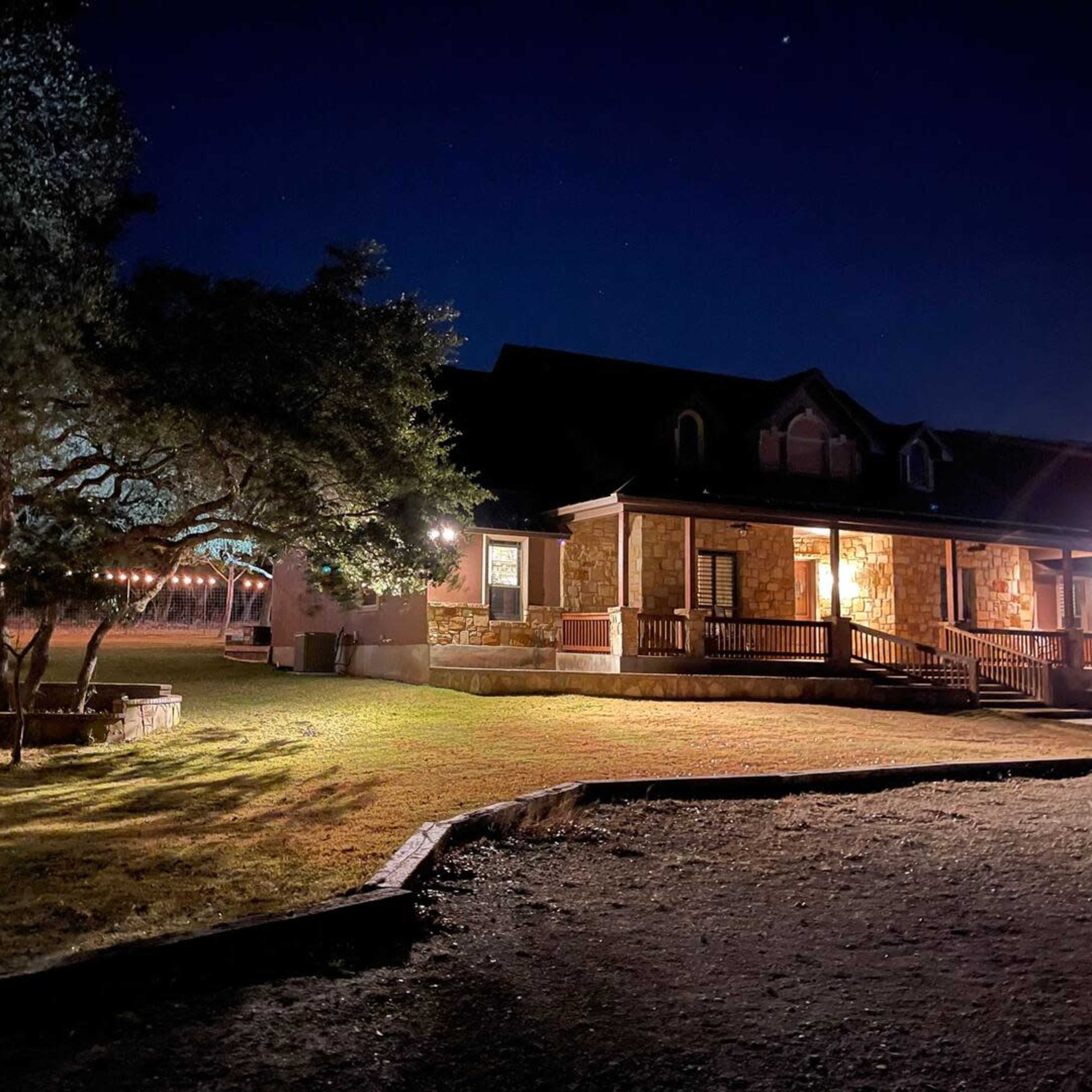A well-lit stone house is surrounded by trees and a grassy yard at night, with decorative string lights illuminating the area.