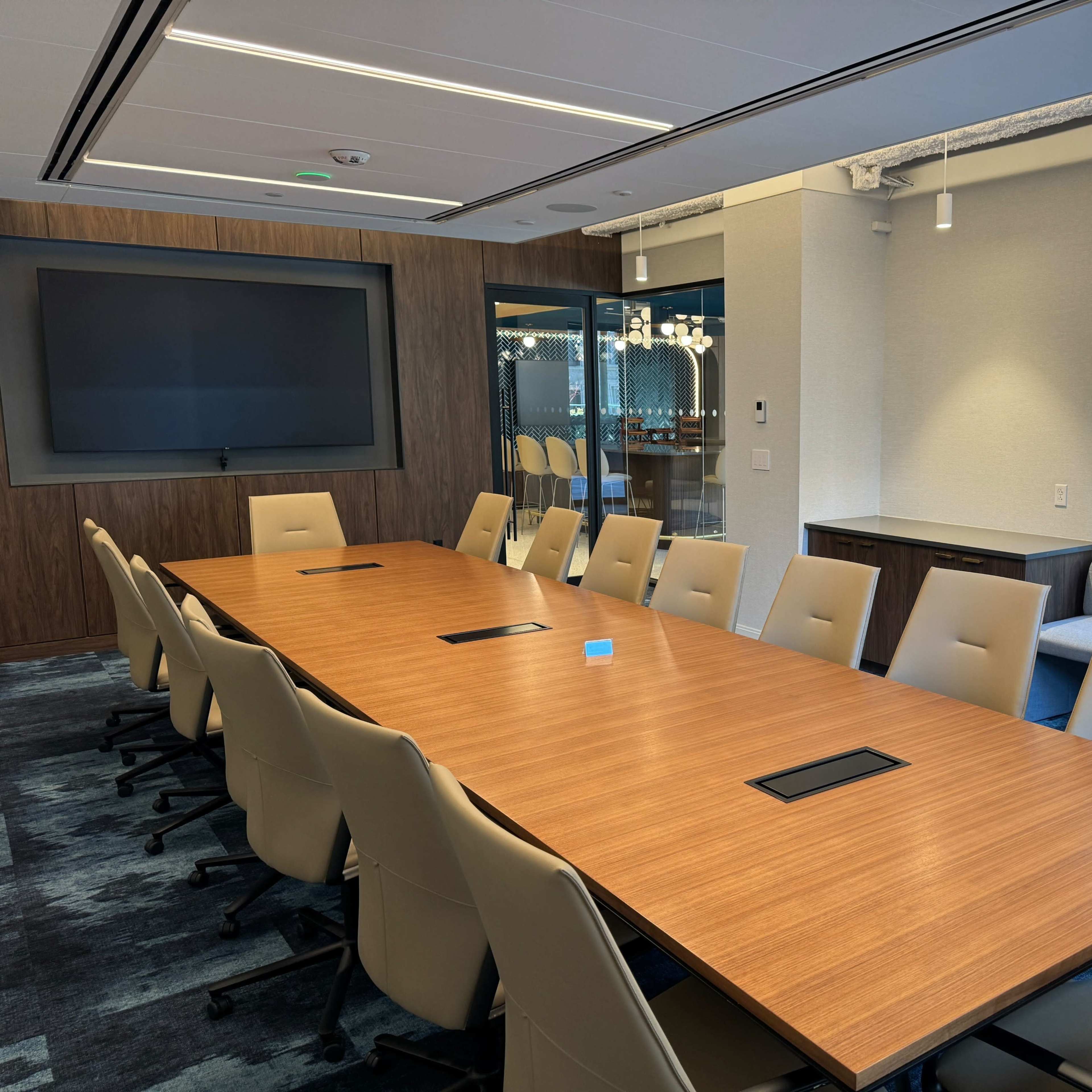 A spacious conference room features a large wooden table surrounded by beige chairs, with a wall-mounted screen and large windows overlooking an outdoor area.