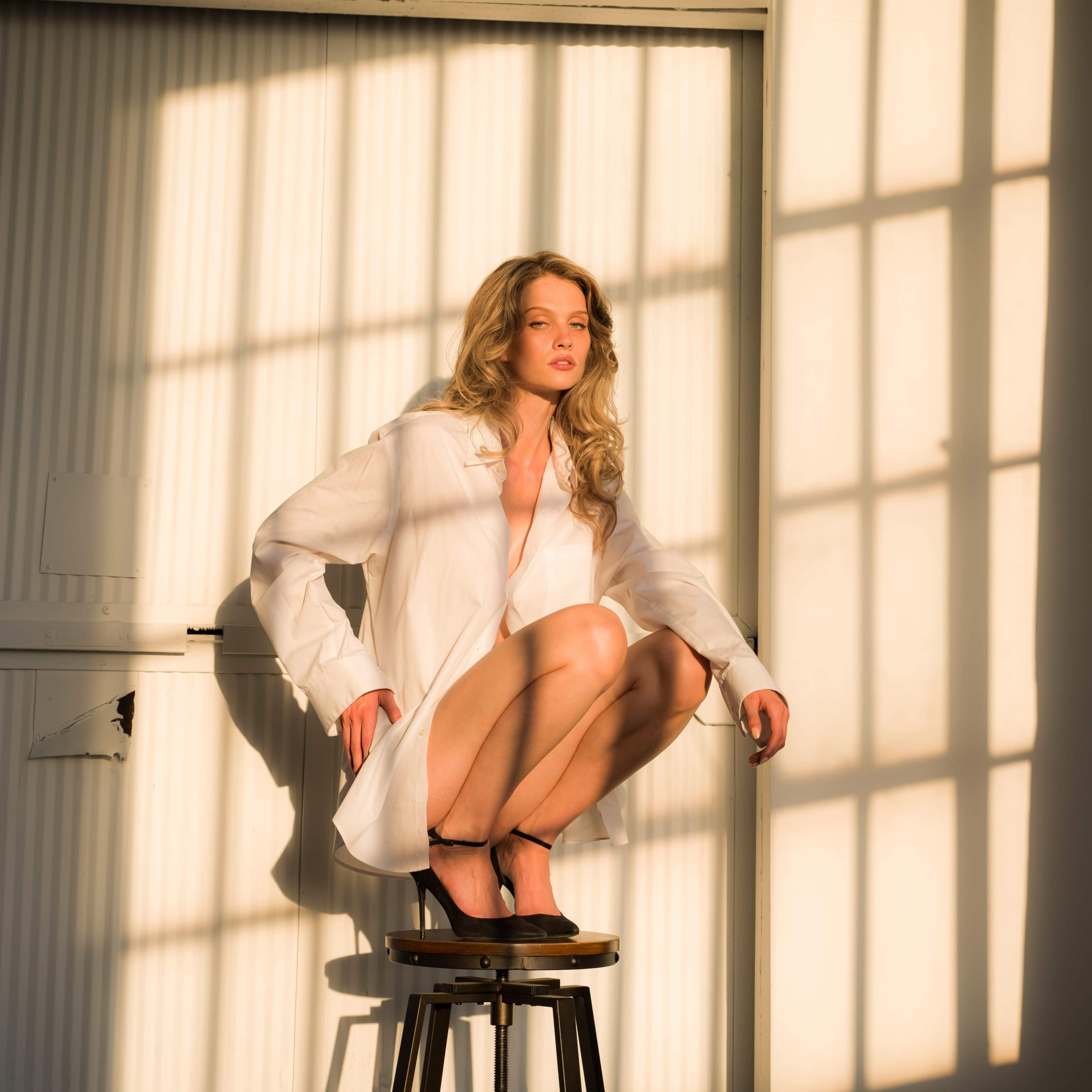 A woman in a white shirt sits on a stool in a well-lit room, casting shadows on the walls.