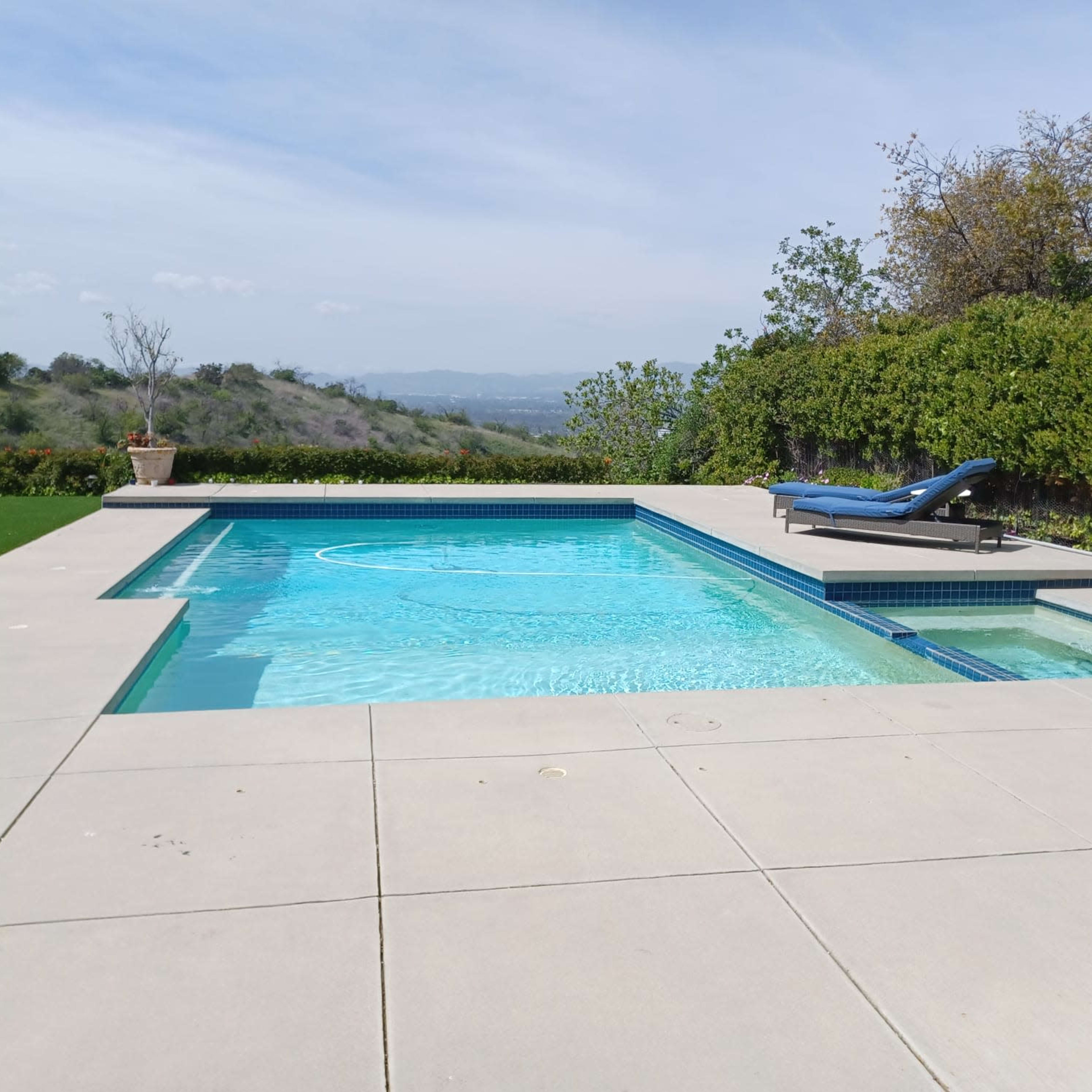A rectangular swimming pool surrounded by a concrete patio, with a lounge chair positioned on one side.